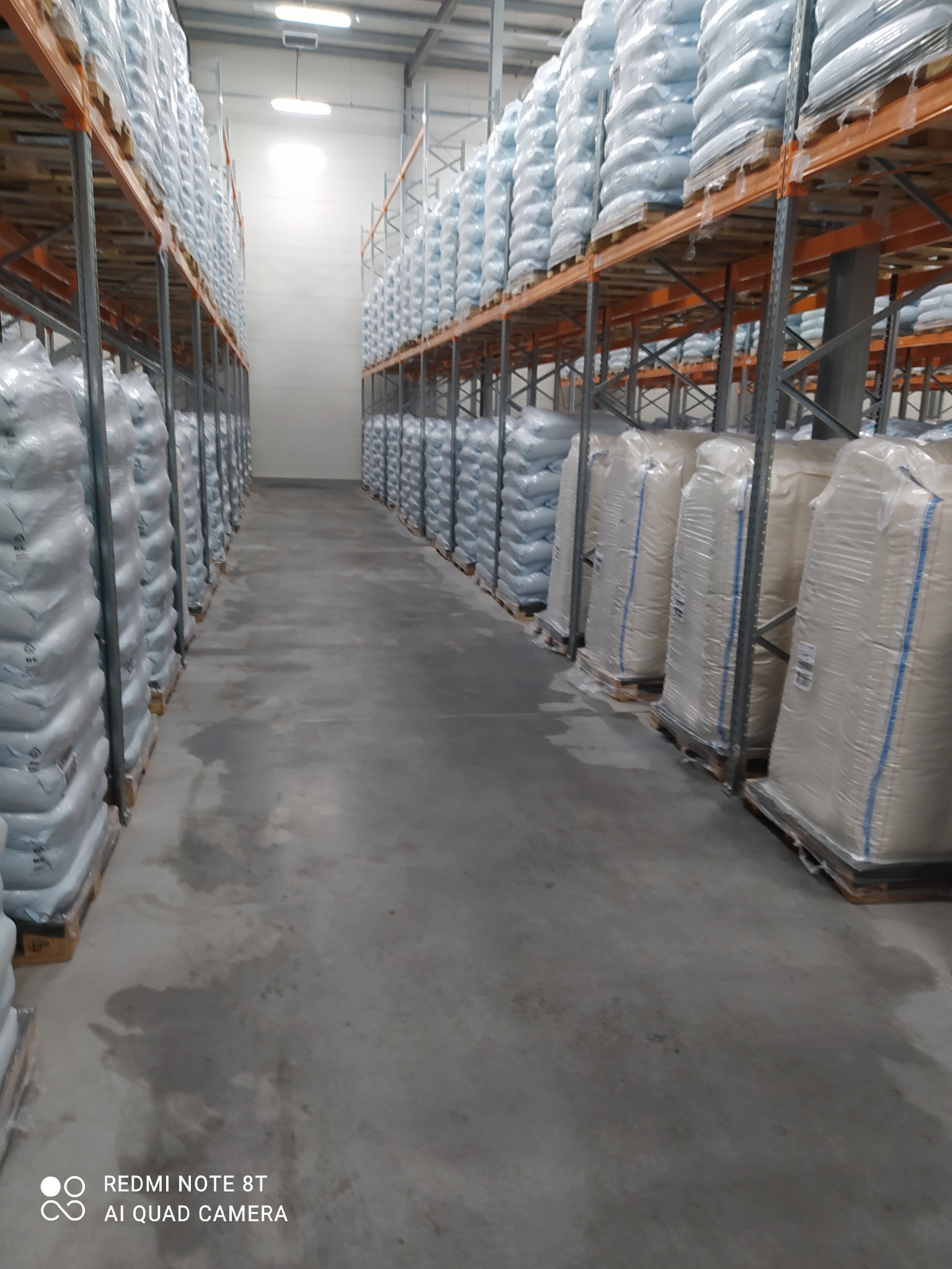 Wnętrze magazynu z rzędami worków składowanych na metalowych regałach, widok z perspektywy podłogi, betonowa posadzka z widocznymi śladami wilgoci.