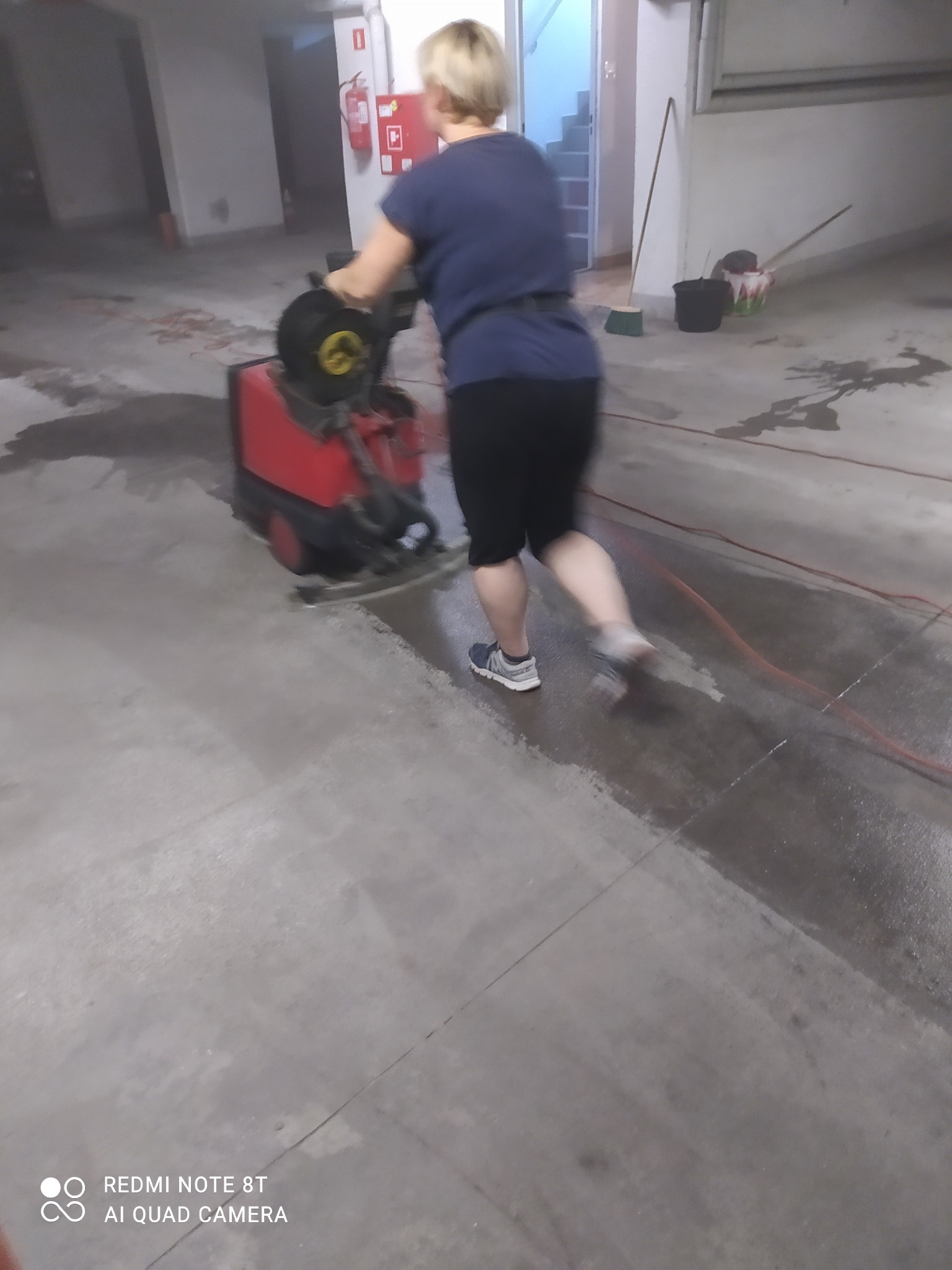 Kobieta obsługuje czerwoną maszynę czyszczącą posadzkę w hali garażowej, widoczne mokre ślady po czyszczeniu, w tle wiadro i miotła.