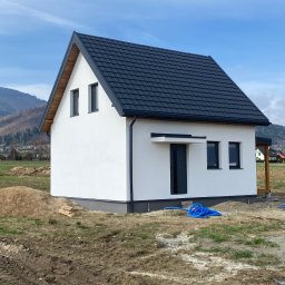 Drev-Const - Niedokończony biały dom z czarnym dachem, widoczne niebieskie węże na błotnistym terenie, góry w tle.