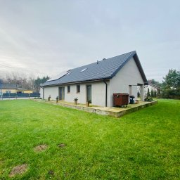 Drev-Const - Nowoczesny dom jednorodzinny z ciemnoszarą dachówką, panelami słonecznymi i zadbanym trawnikiem, widok z perspektywy ogrodu w pochmurny dzień.