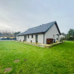 Nowoczesny dom jednorodzinny z ciemnoszarą dachówką, panelami słonecznymi i zadbanym trawnikiem, widok z perspektywy ogrodu w pochmurny dzień.