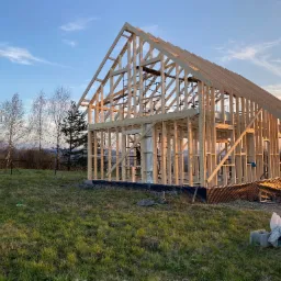 Surowy szkielet domu w trakcie budowy, widoczny na tle zachodzącego słońca i krajobrazu z drzewami, z elementami izolacji i fundamentami.