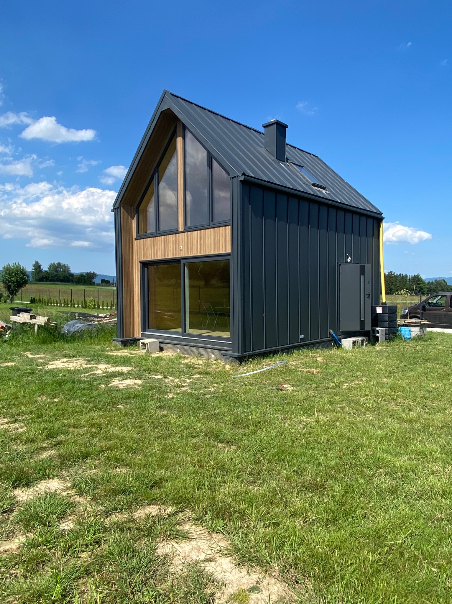 Nowoczesny, czarny dom z metalowym dachem i drewnianą fasadą, duży przeszklenia, trawnik wokół, błękitne niebo.