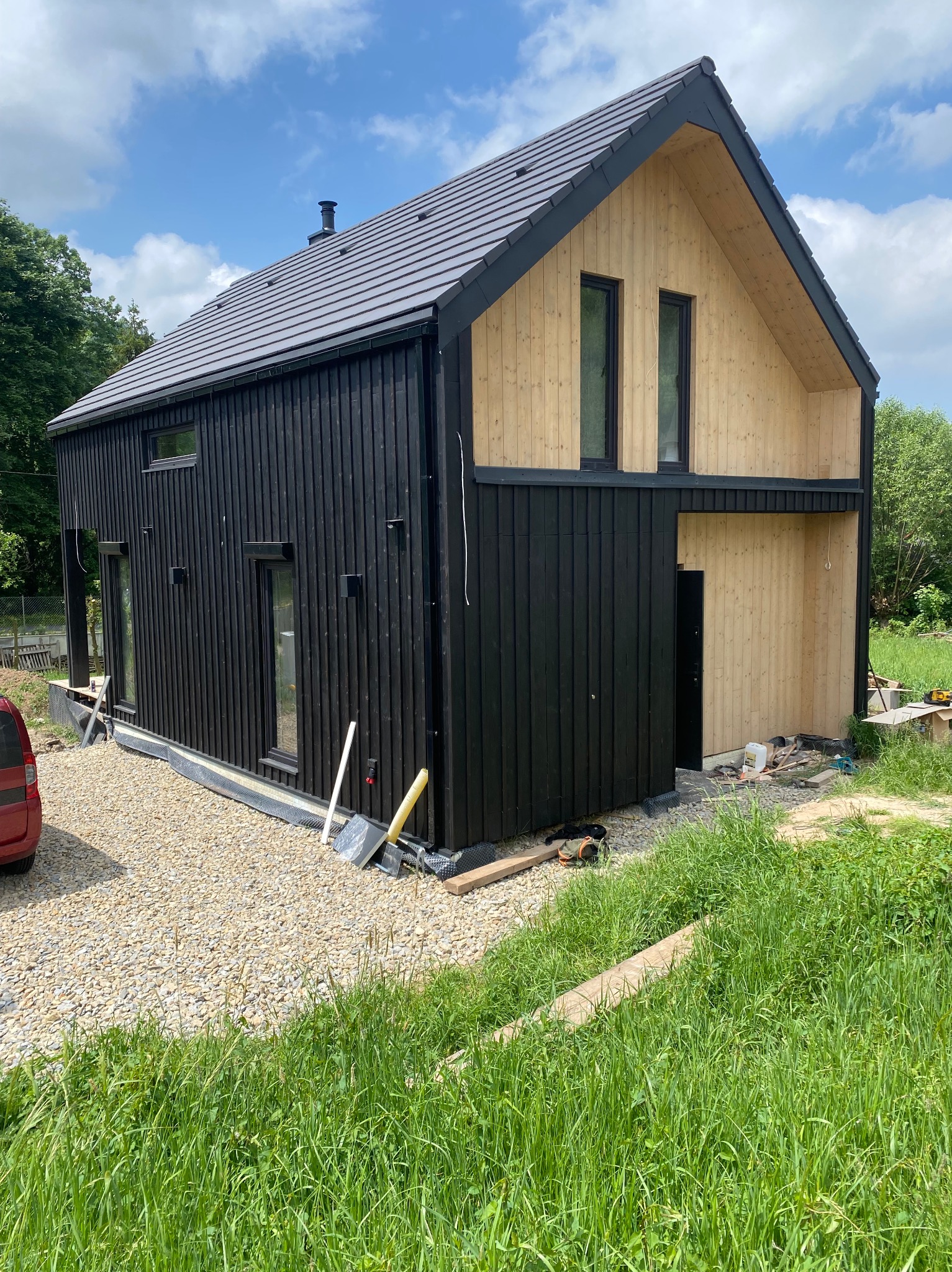 Nowoczesny dom z drewnianą elewacją w kolorze czarnym i naturalnym, z szarą dachówką, na etapie wykończenia, widoczne narzędzia i materiały budowlane.