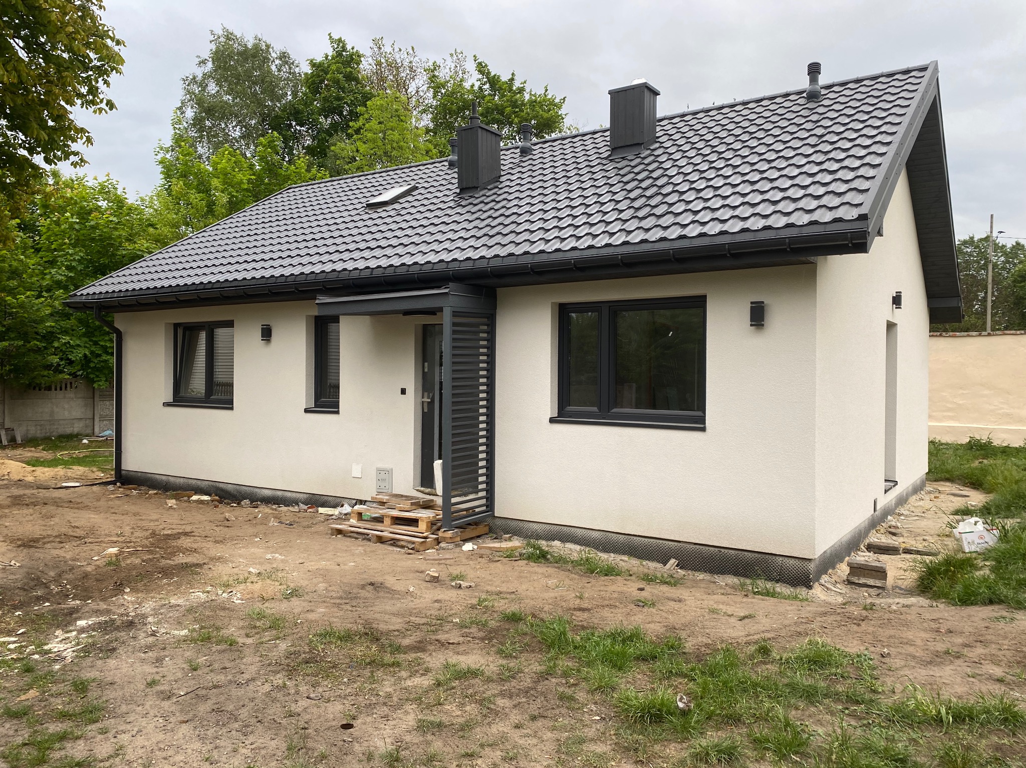 Nowoczesny, parterowy dom z białym tynkiem i ciemnoszarą dachówką, widoczne palety przy wejściu, teren wokół domu w trakcie prac wykończeniowych.