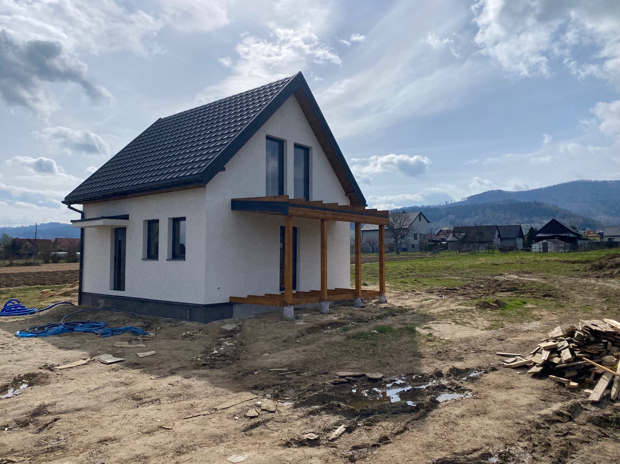 Nowoczesny dom szkieletowy z ciemnym dachem i drewnianą konstrukcją werandy w trakcie budowy, widoczne materiały budowlane i błoto na działce.