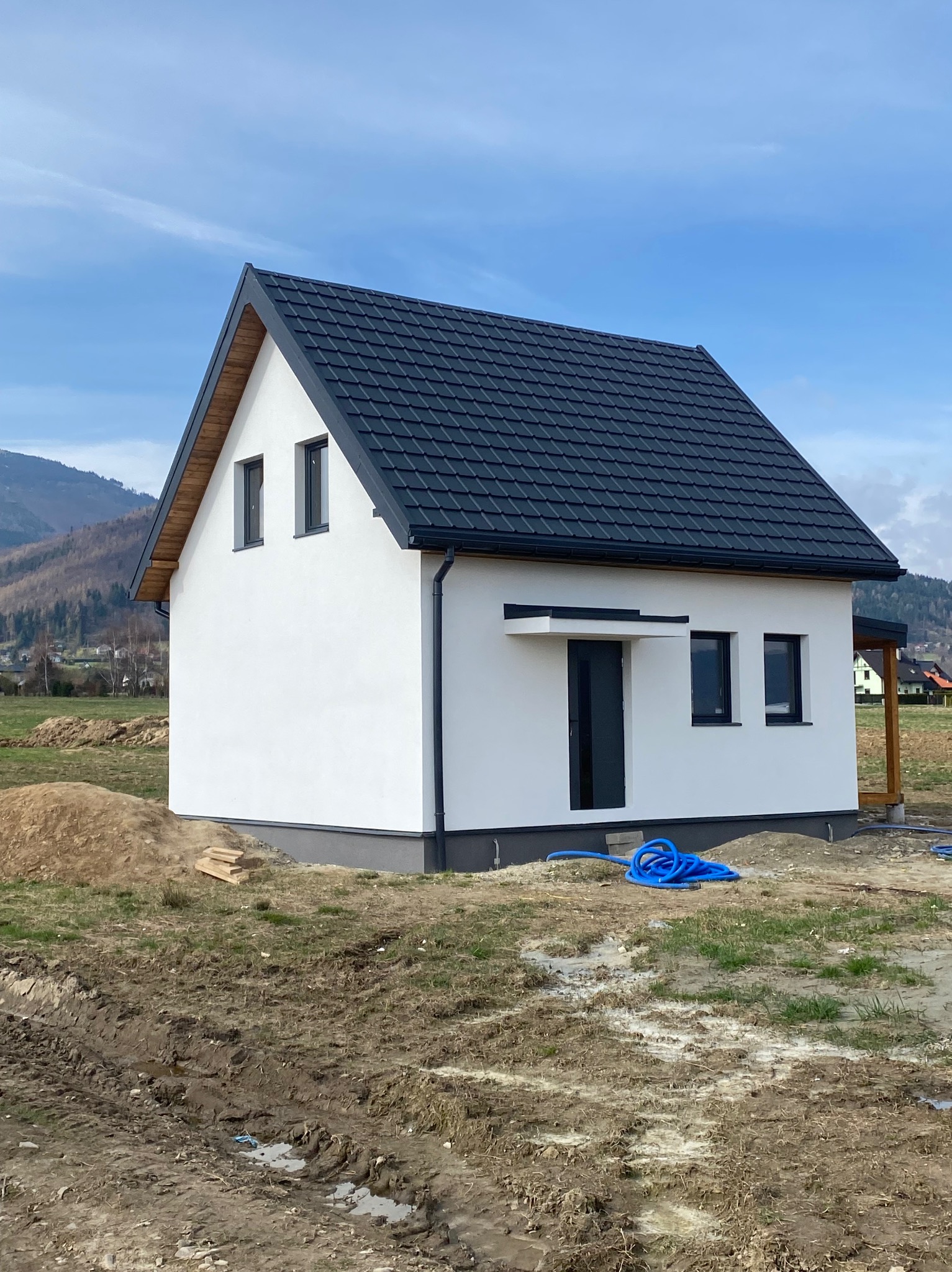 Niedokończony biały dom z czarnym dachem, widoczne niebieskie węże na błotnistym terenie, góry w tle.