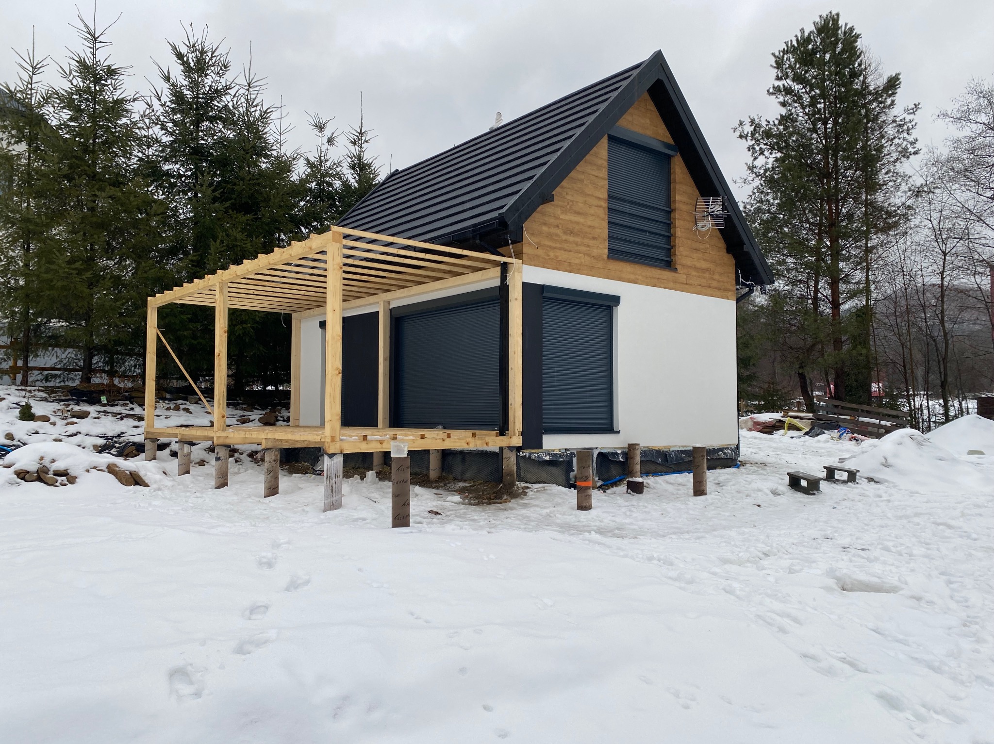 Niedokończony dom z drewnianym szkieletem w zimowym krajobrazie, widoczne drewniane belki i białe ściany, czarny dach z blachy trapezowej i grafitowe rolety zewnętrzne.