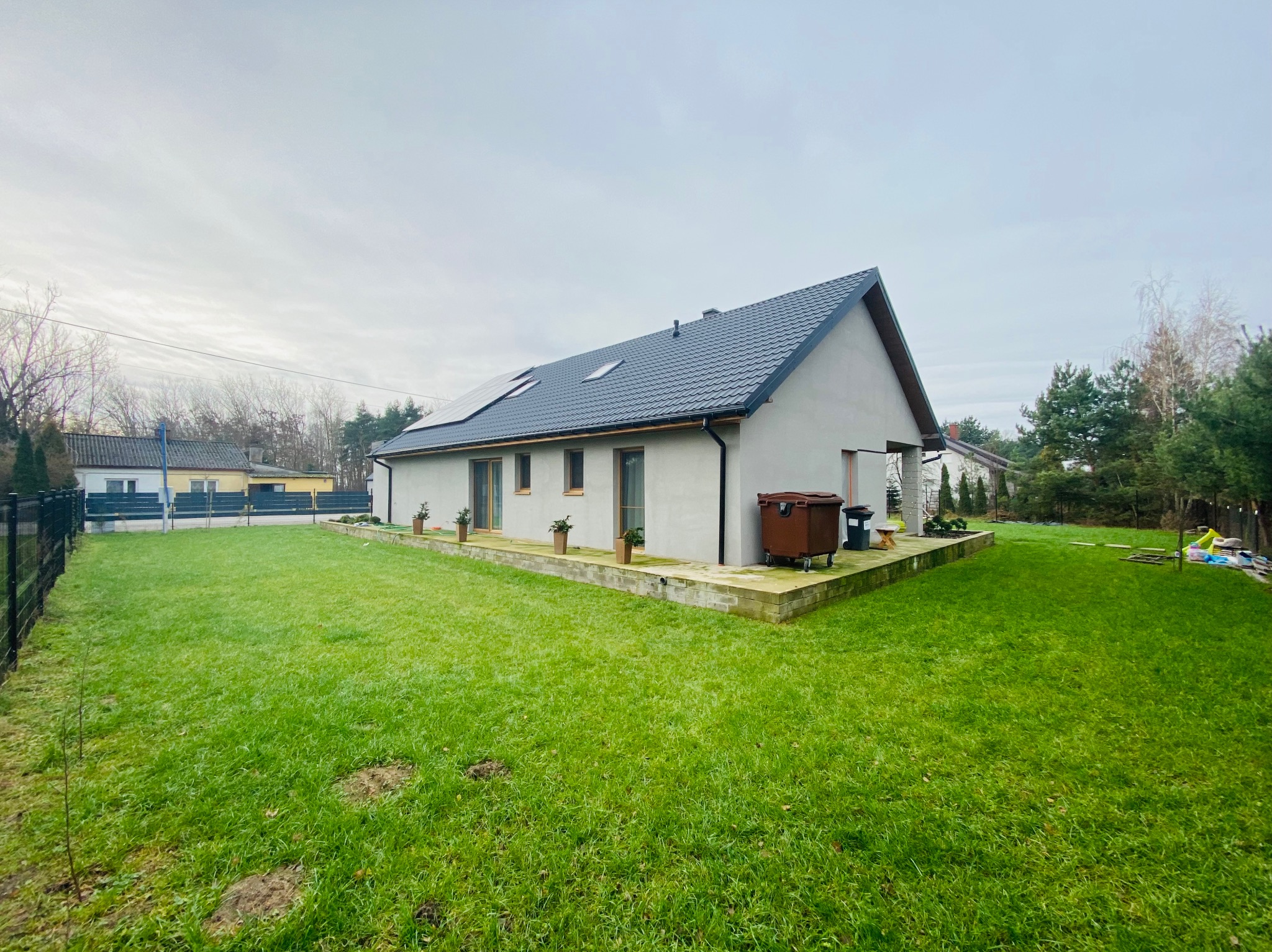 Nowoczesny dom jednorodzinny z ciemnoszarą dachówką, panelami słonecznymi i zadbanym trawnikiem, widok z perspektywy ogrodu w pochmurny dzień.