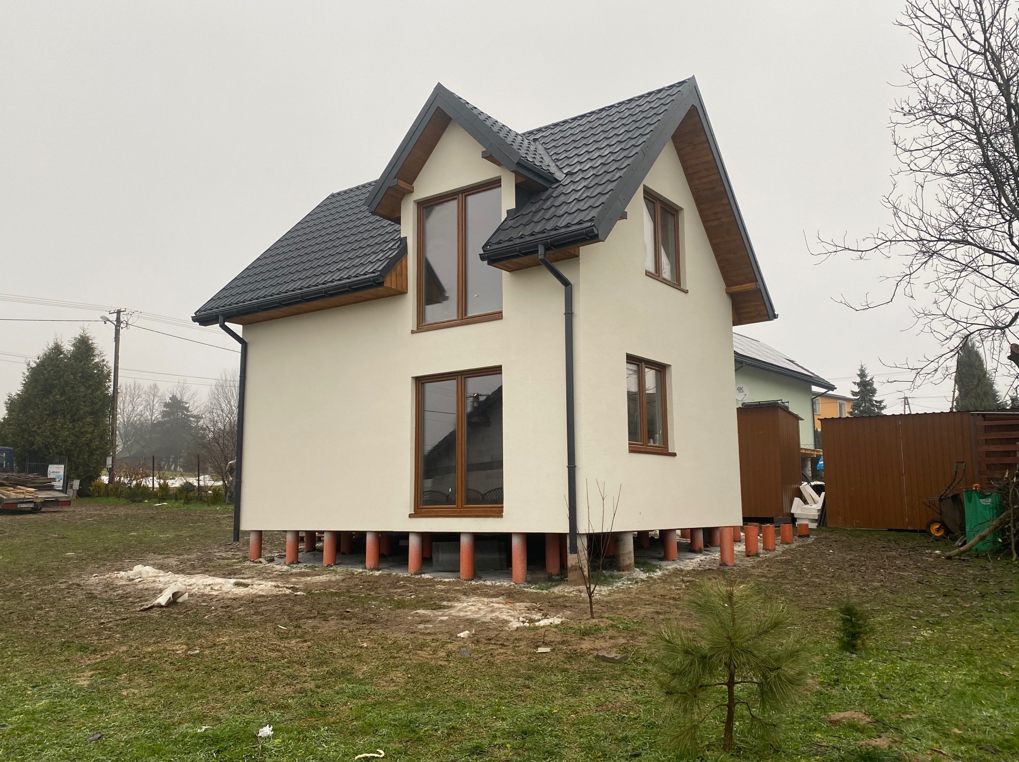 Nowo wybudowany, nieotynkowany dom szkieletowy na podwyższeniu z pomalowanymi na pomarańczowo słupami, z ciemnym dachem i brązowymi ramami okiennymi w pochmurny dzień.