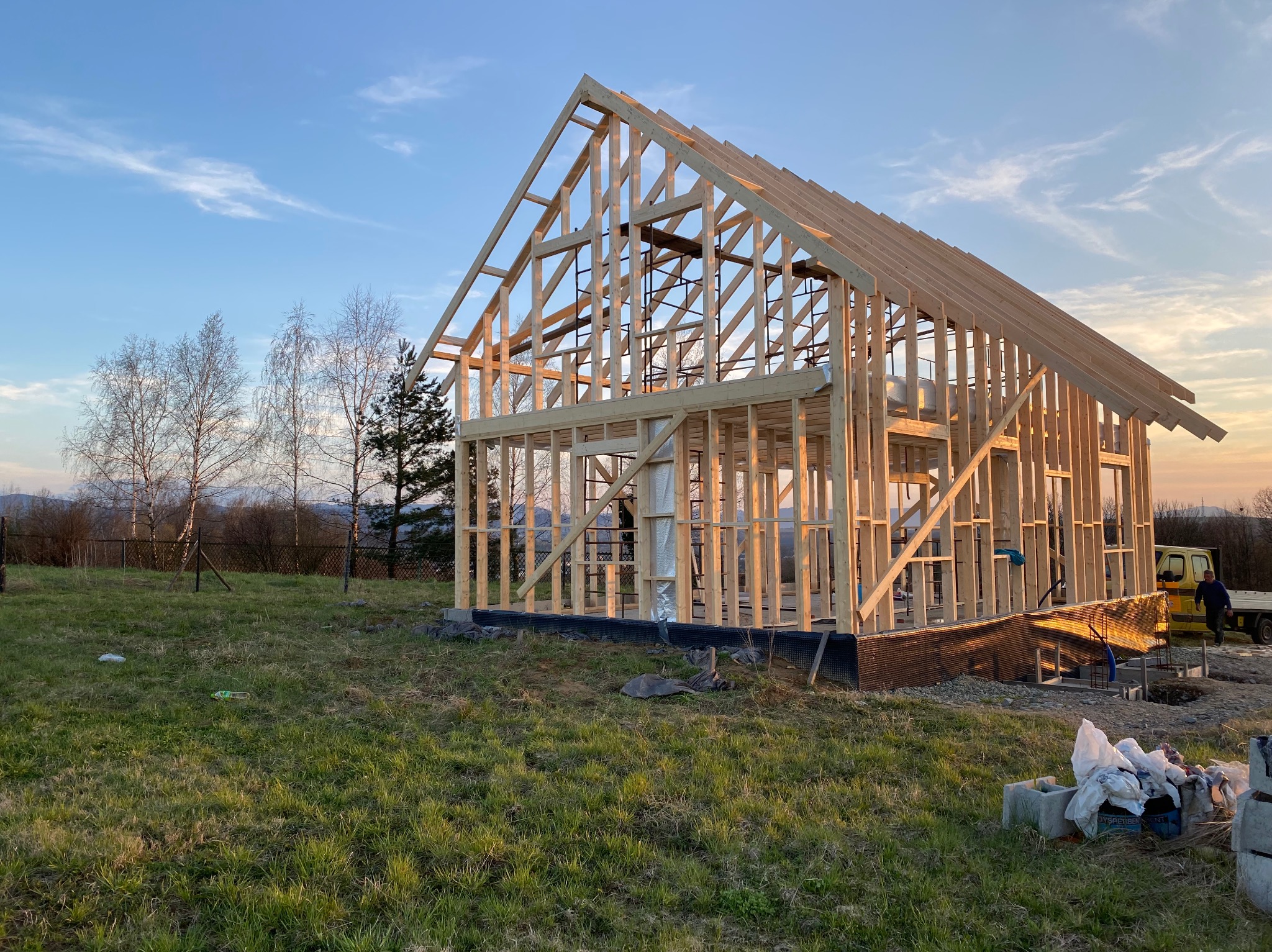 Surowy szkielet domu w trakcie budowy, widoczny na tle zachodzącego słońca i krajobrazu z drzewami, z elementami izolacji i fundamentami.
