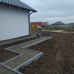 MARAS - Krawężniki wyznaczające ścieżkę wypełnioną drobnym żwirem, ułożone w zygzakowaty wzór obok białego domu z ciemnym dachem, w tle widoczny żółty samochód dostawczy i budowa domu w stanie surowym.