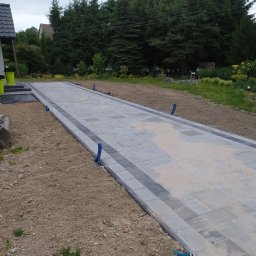 MARAS - Ułożona aleja z kostki brukowej w odcieniach szarości z ciemniejszym obramowaniem, widoczne niebieskie rurki instalacyjne wzdłuż krawędzi, teren wokół jeszcze niezagospodarowany, w tle iglaki.