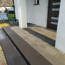 Układanie kostki brukowej w Polanicy-Zdrój: widok na świeżo ułożony fragment schodów wejściowych i chodnika z kostki w dwóch odcieniach szarości, z widocznymi narzędziami i materiałami budowlanymi...