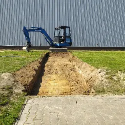 Niebieska minikoparka Hitachi EX30 stoi obok wykopu w ziemi, na tle szarej hali z blachy falistej i zielonego trawnika.