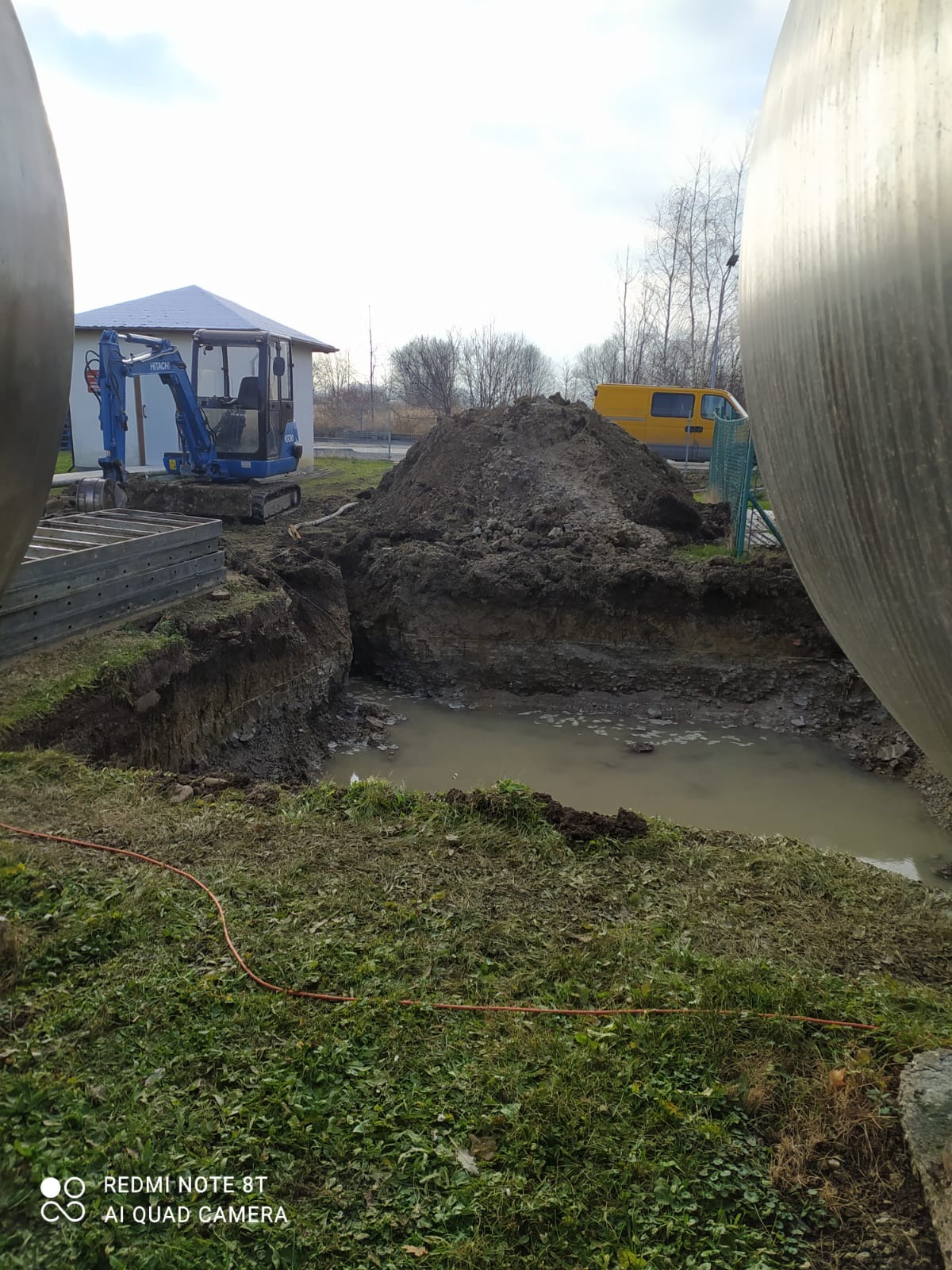 Wykop wypełniony wodą, obok sterta ziemi, mała koparka Hitachi i żółty bus w tle, widok z poziomu gruntu.