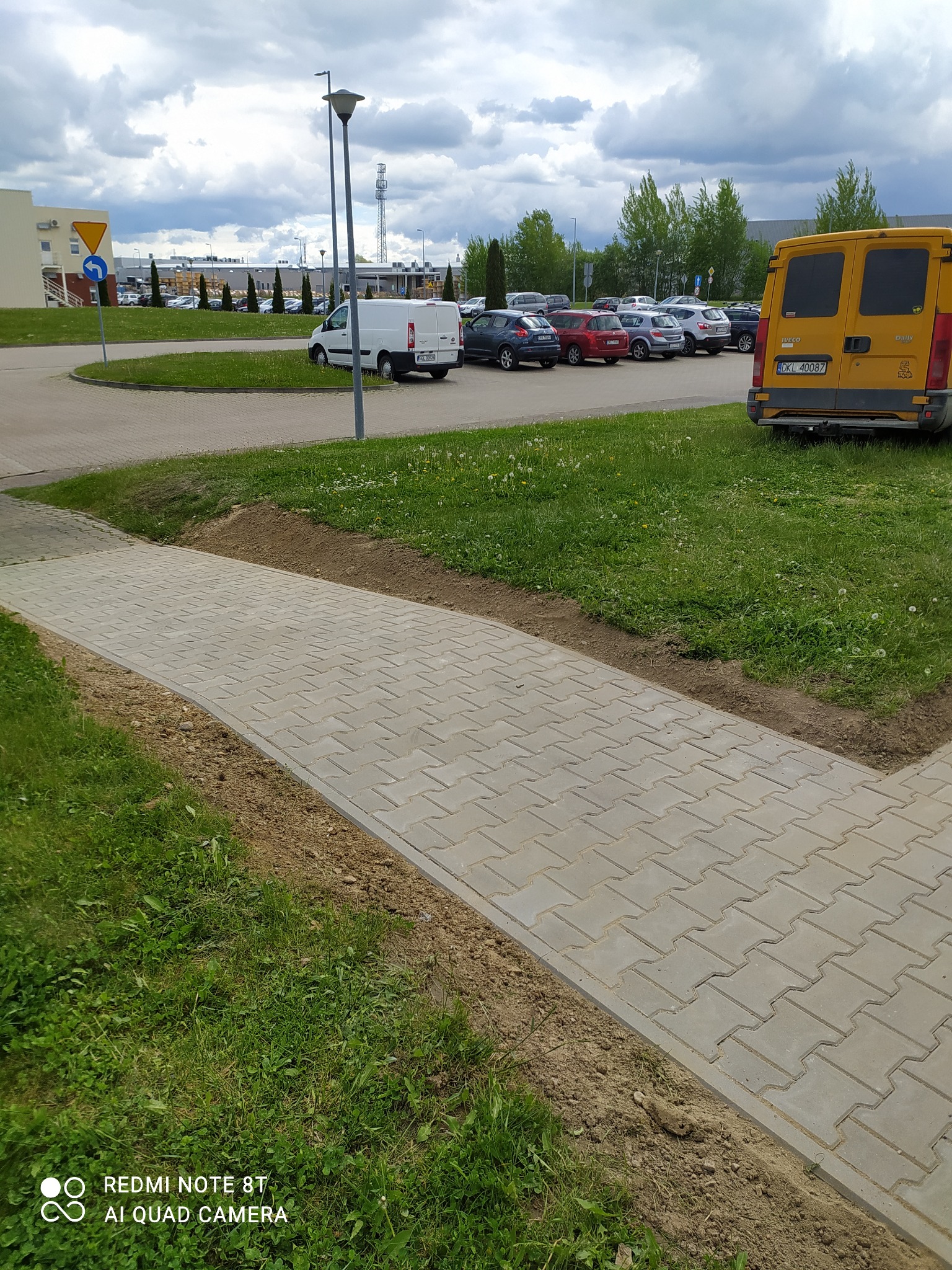 Nowo ułożony chodnik z szarej kostki brukowej na tle trawnika, z widocznym fragmentem żółtego samochodu dostawczego i parkingu w oddali.