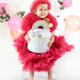 Uśmiechnięta dziewczynka w różowej tiulowej sukience i opasce, siedzi na białym krześle trzymając tort z tęczą i lizakami na białym tle z balonami.