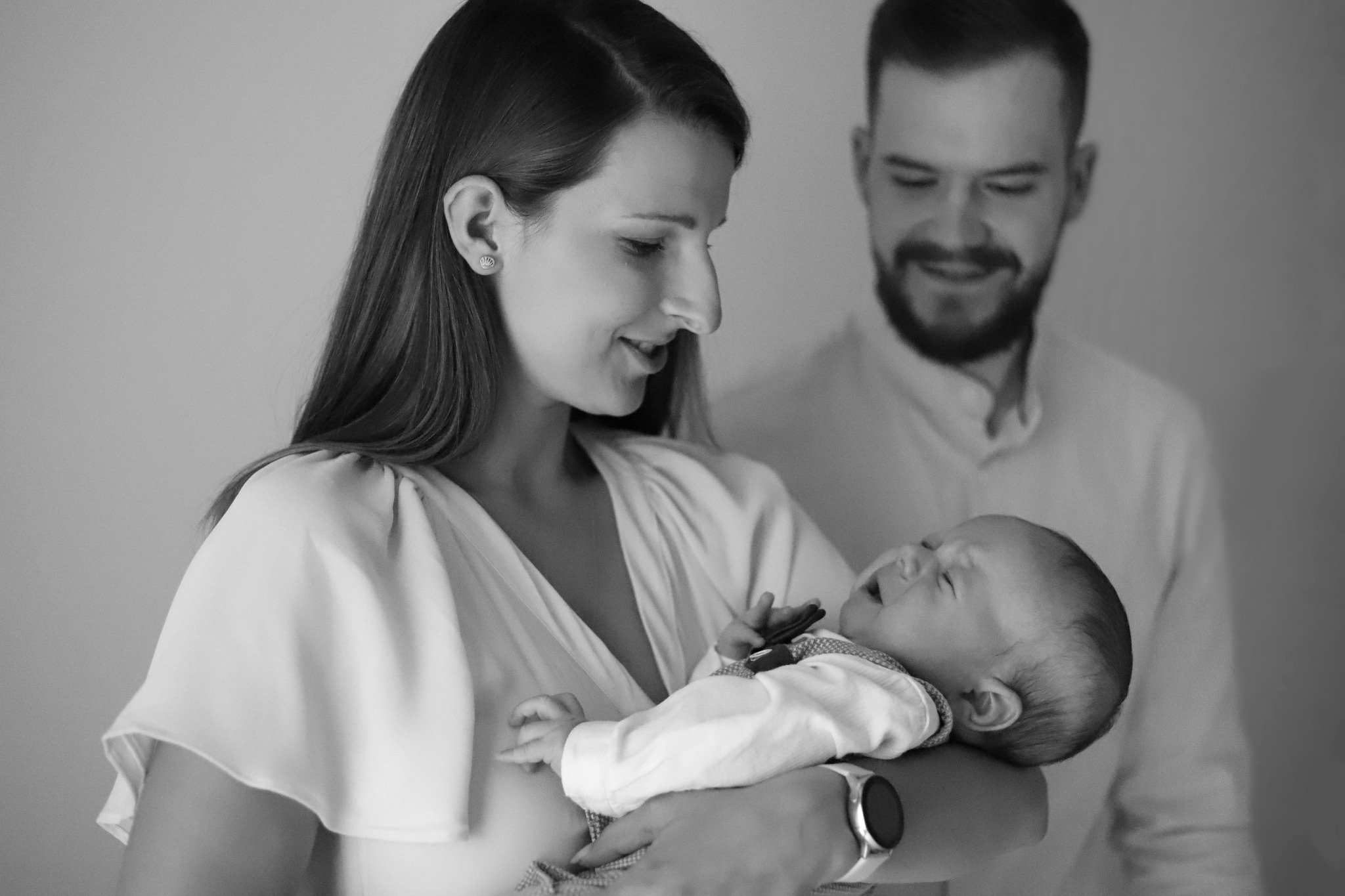 Czarno-białe zdjęcie przedstawia młodą mamę trzymającą na rękach noworodka, obok stoi uśmiechnięty tata. Mama ma długie włosy i kolczyki. Dziecko ma na sobie koszulę w kratę i ciemną muchę.