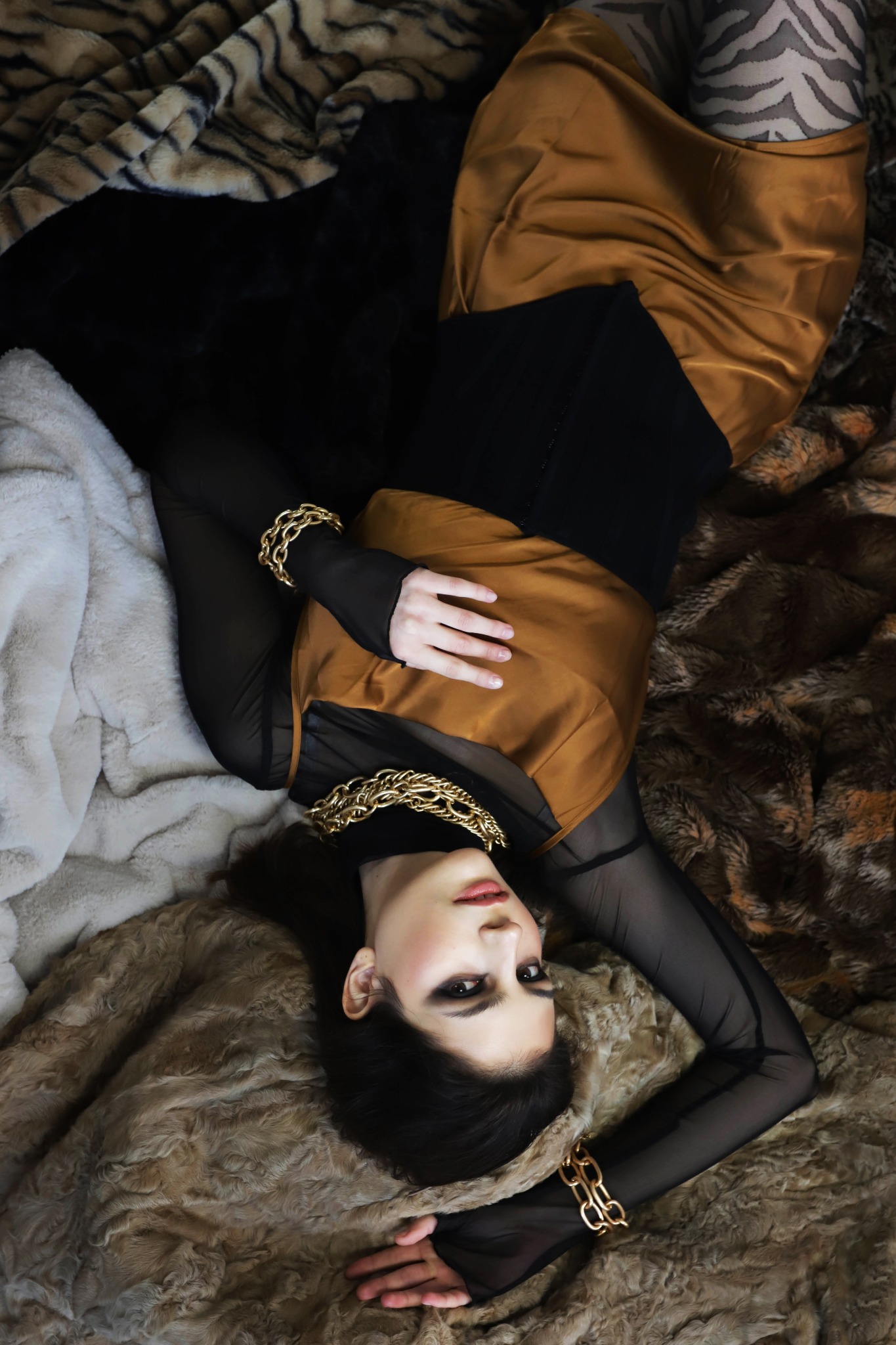 Kobieta w złotej sukience i czarnej bluzce z siateczki, leżąca na futrzanym tle z motywami zwierzęcymi, ozdobiona grubym złotym łańcuchem na szyi i bransoletkami.