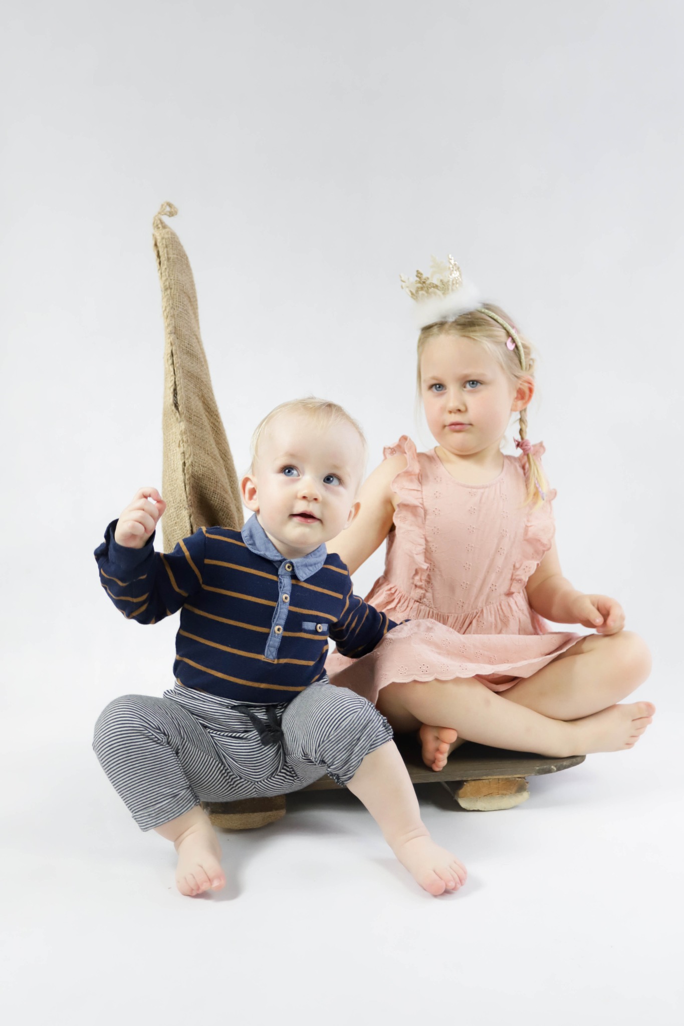 Dwoje dzieci, chłopiec w pasiastym ubranku i dziewczynka z koroną na głowie w różowej sukience, siedzą na drewnianej desce na tle białego studia z dekoracyjnym stożkiem z juty.