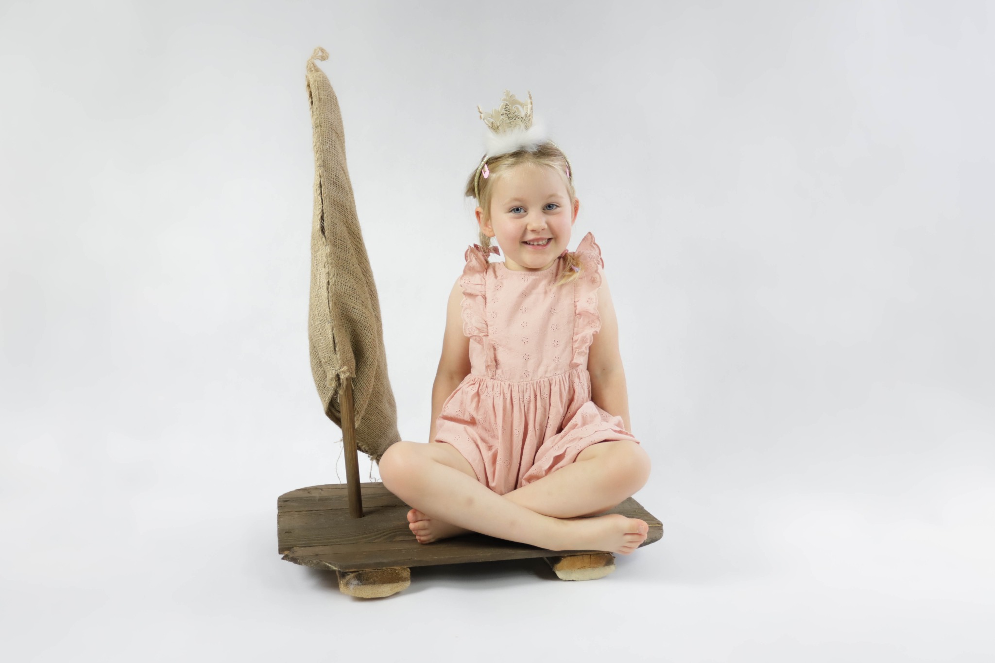 Uśmiechnięta dziewczynka w koronie i różowej sukience siedzi na drewnianej platformie z rekwizytem żagla z juty na białym tle.
