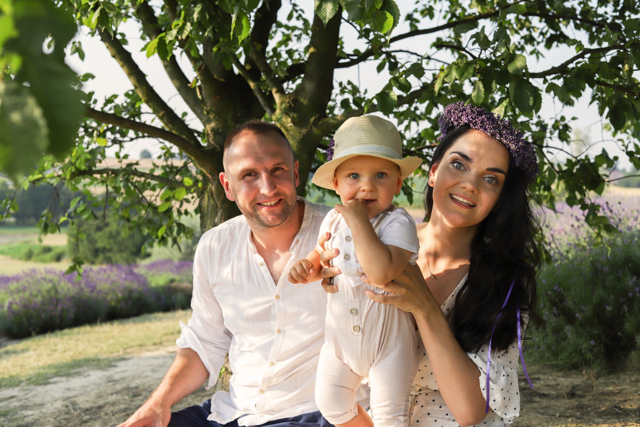 Uśmiechnięta rodzina: ojciec siedzący obok drzewa, matka z wiankiem z lawendy na głowie trzymająca na rękach roczne dziecko w kapeluszu, lawendowe pole w tle.