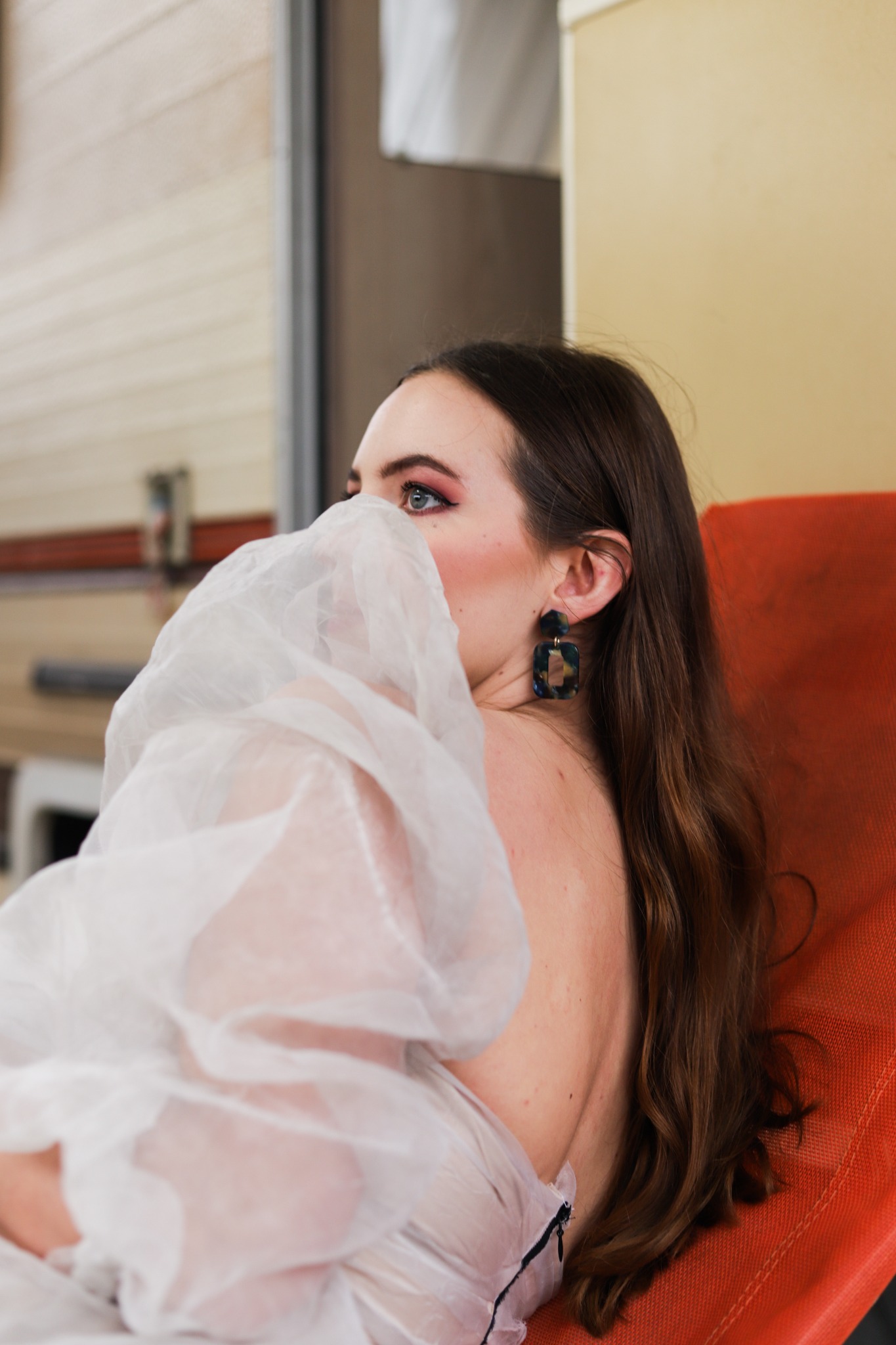 Portret kobiety z rozwianym białym materiałem zakrywającym część twarzy, widoczne oko z różowym makijażem, ciemne włosy i duże, geometryczne kolczyki na pomarańczowym krześle.