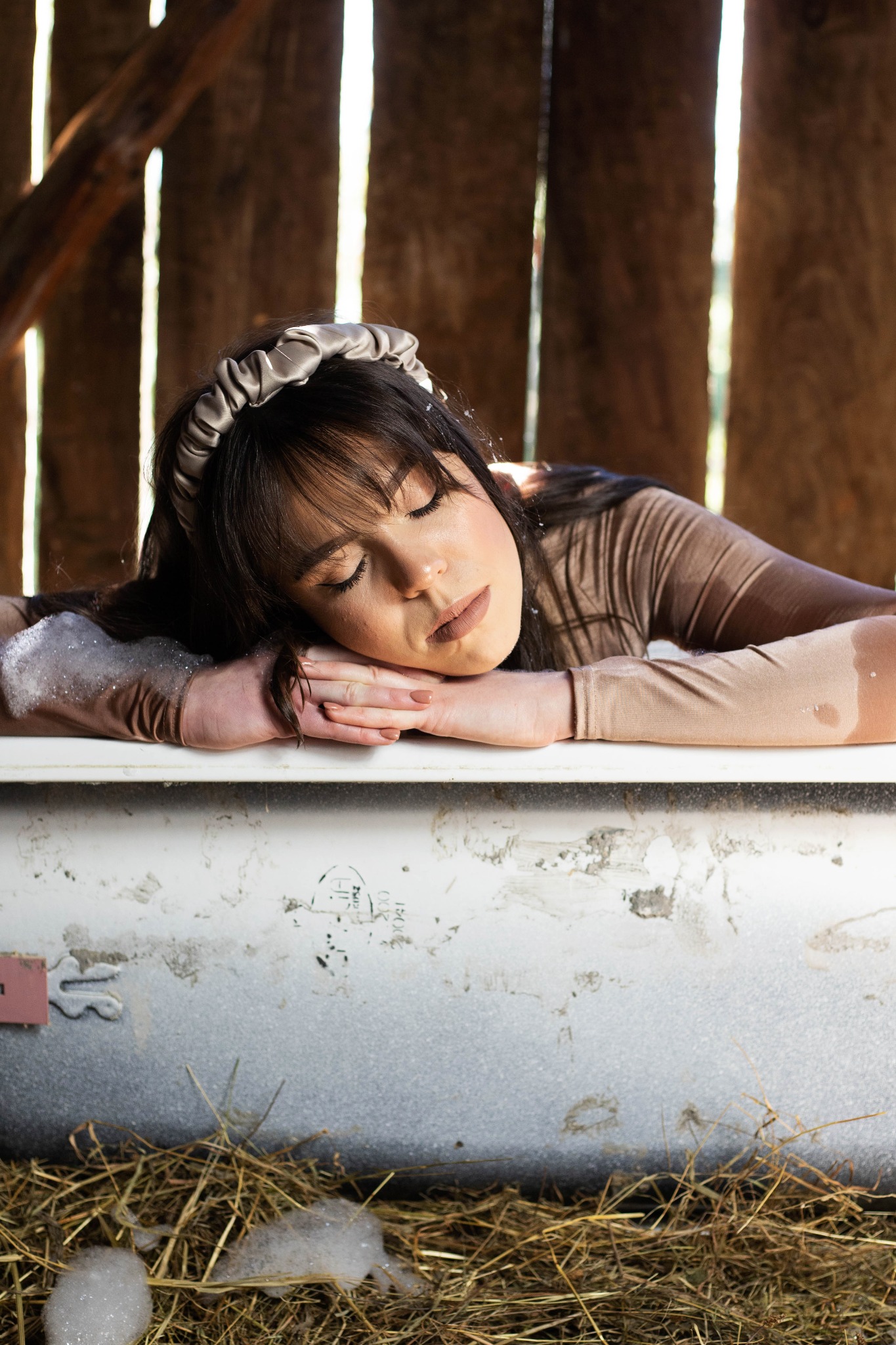 Kobieta z zamkniętymi oczami, z satynową opaską na włosach, opiera się o brzeg starej wanny wypełnionej pianą, na tle drewnianej ściany stodoły i siana.