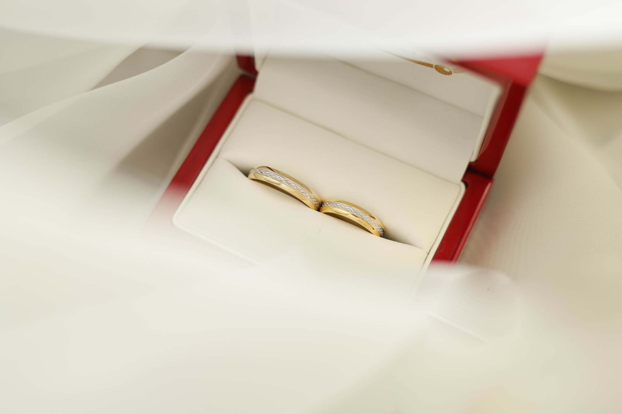 Dwie złote obrączki z delikatnym zdobieniem w czerwonym pudełku na białym, delikatnym materiale.