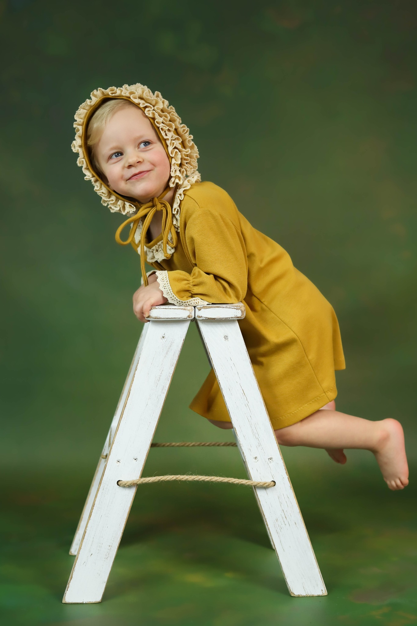 Uśmiechnięta blond dziewczynka w musztardowej sukience i czepku z falbankami, oparta o białą, drewnianą drabinkę na tle zielonego płótna.