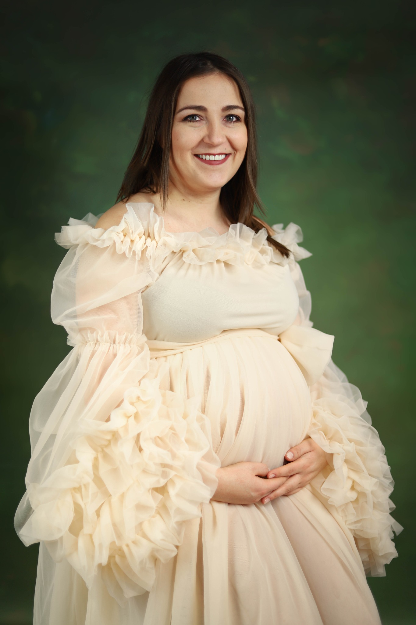 Uśmiechnięta kobieta w ciąży, w kremowej, zwiewnej sukni z falbanami, trzyma dłonie na brzuchu na tle zielonego, malowanego tła.
