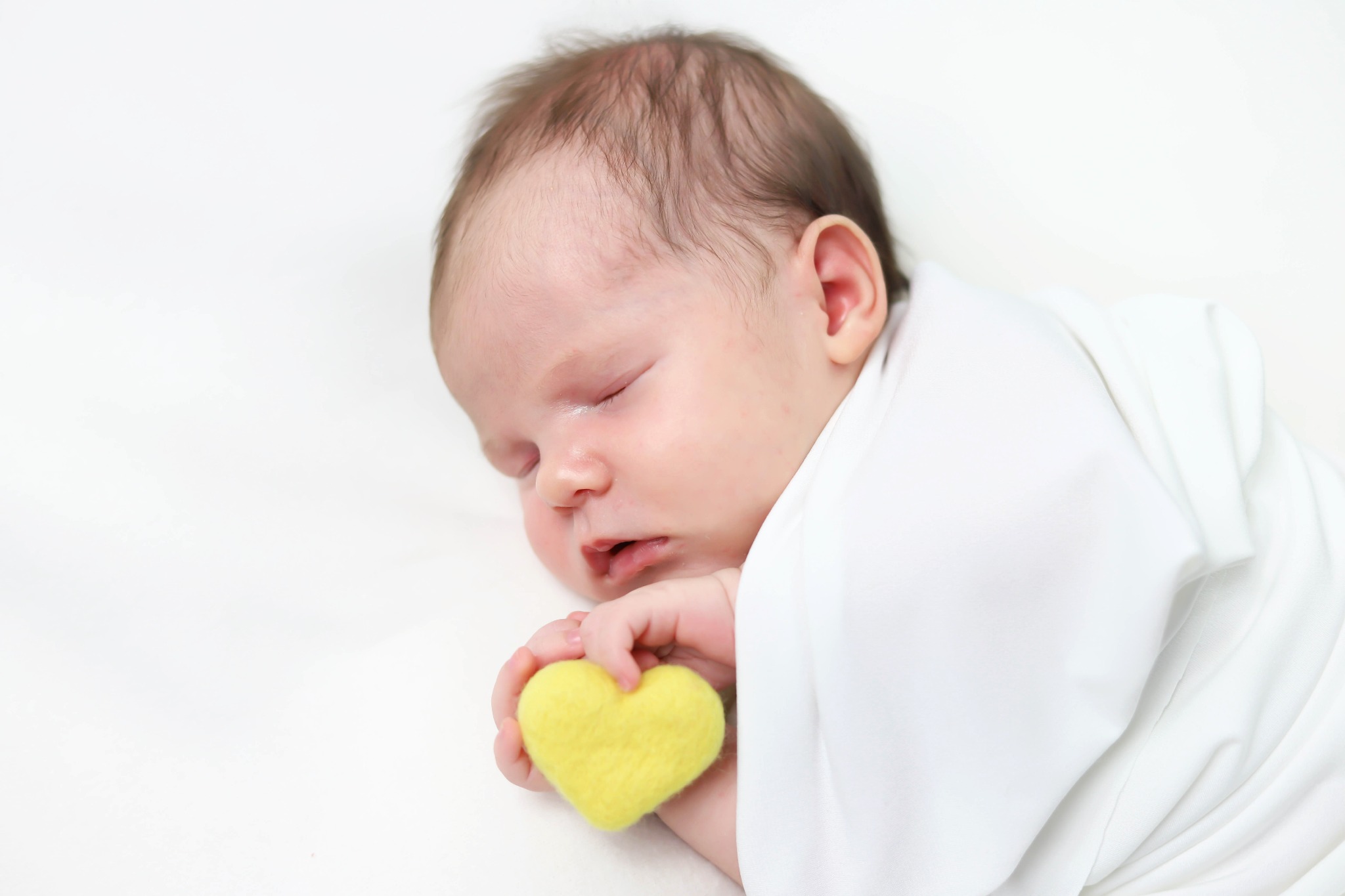 Śpiący noworodek owinięty w białe płótno, trzymający w dłoni filcowe żółte serce na białym tle.