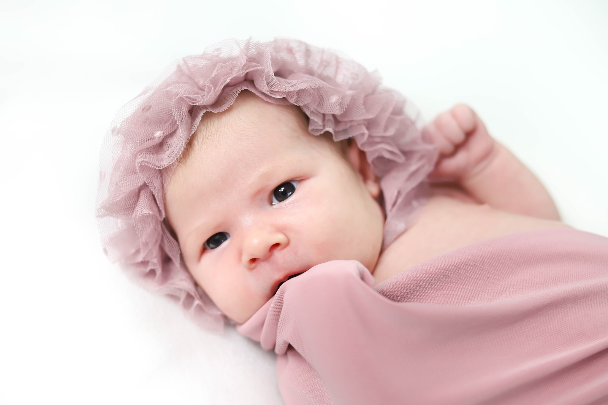 Noworodek w tiulowym czepku i różowym otulaczu na białym tle, z wyraźnym spojrzeniem w obiektyw.