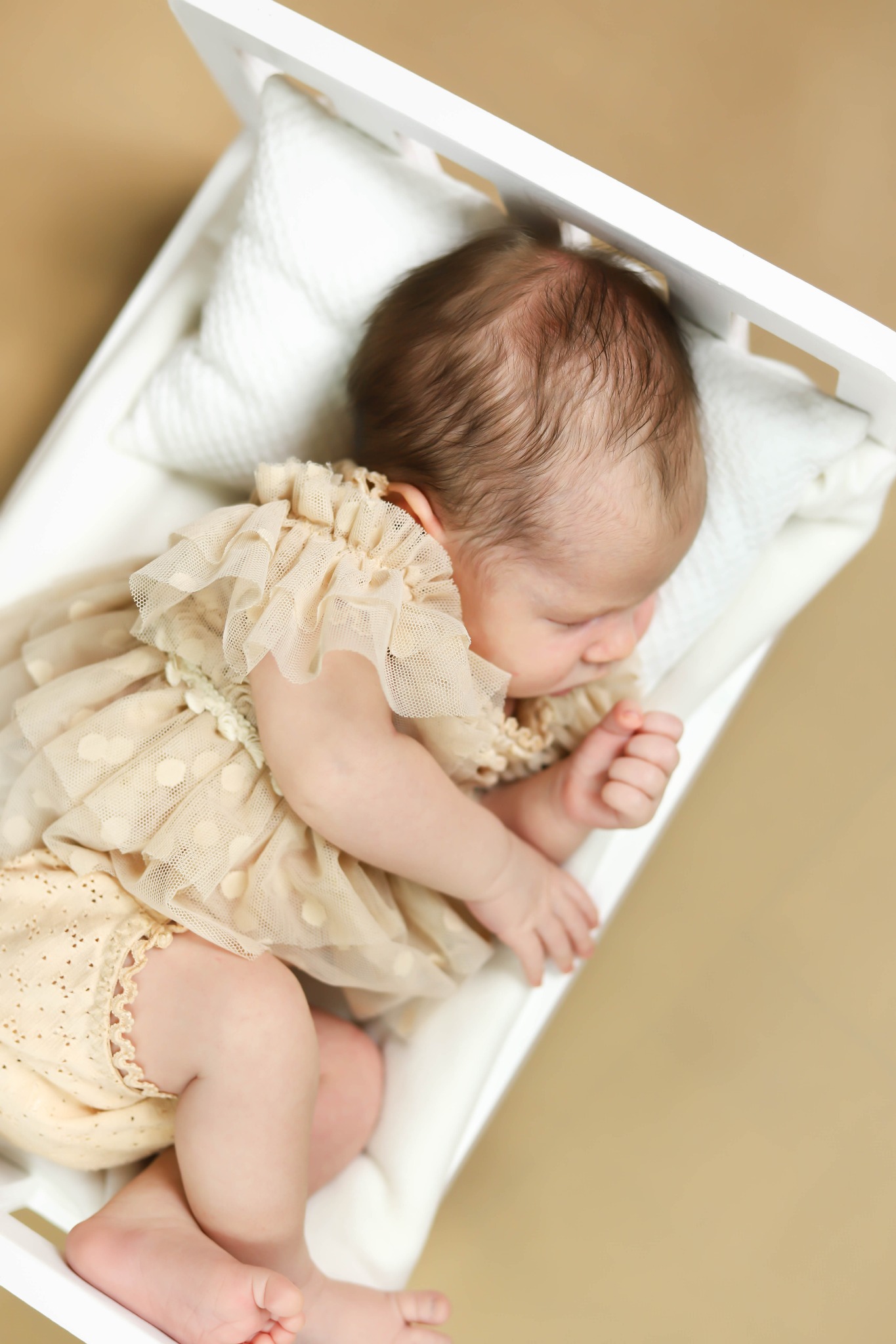 Śpiący noworodek w beżowej sukience z falbankami, leżący w małym białym łóżeczku na beżowym tle, widok z góry.
