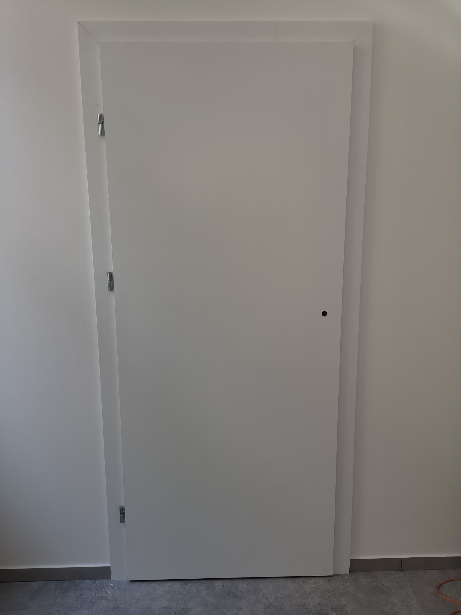 Białe, minimalistyczne drzwi wewnętrzne z ukrytymi zawiasami, zamontowane w białej ścianie na szarym podłożu.