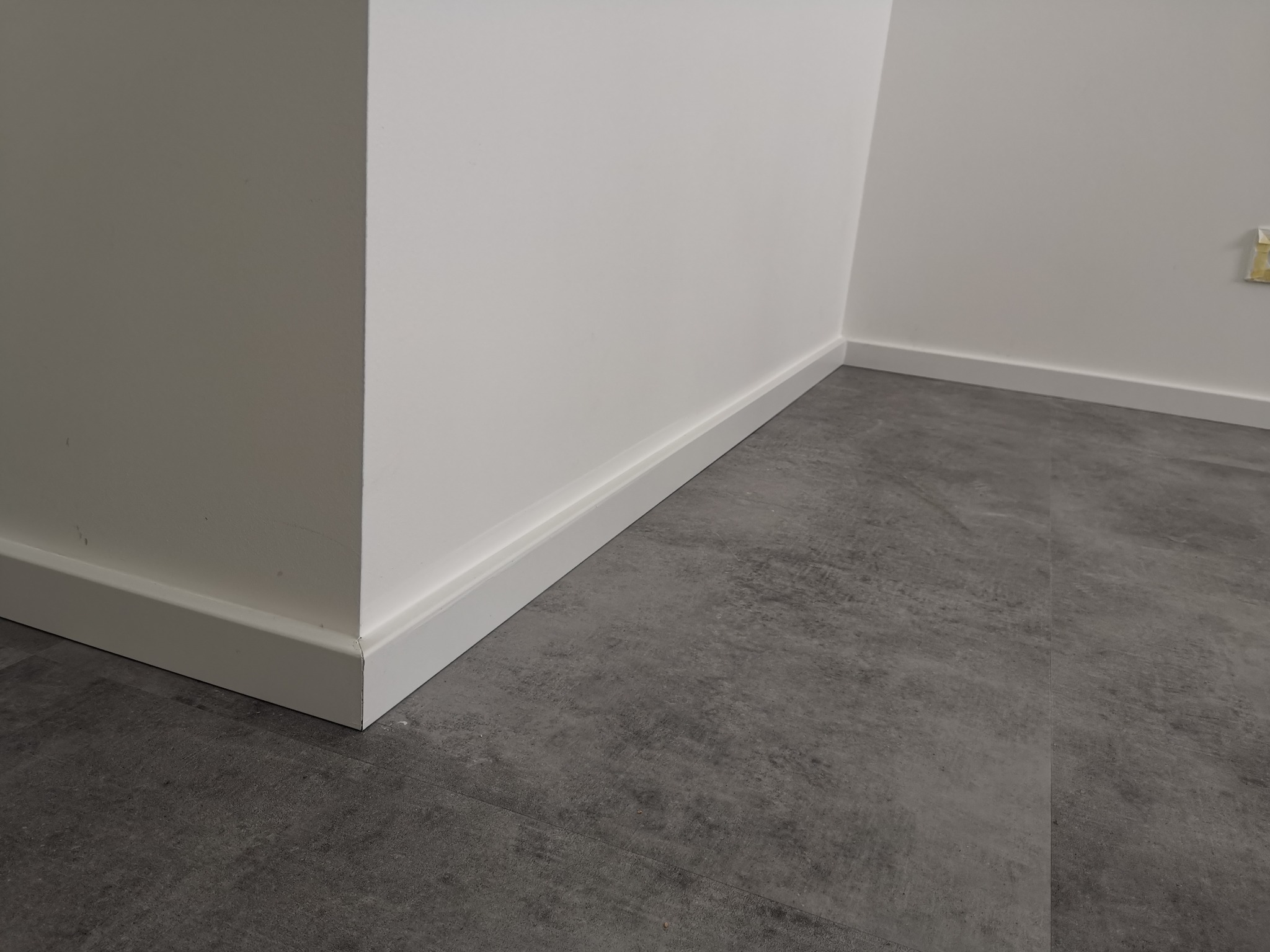 Szare panele podłogowe imitujące beton, białe listwy przypodłogowe przy ścianie pomalowanej na biało, widoczny fragment gniazdka elektrycznego.