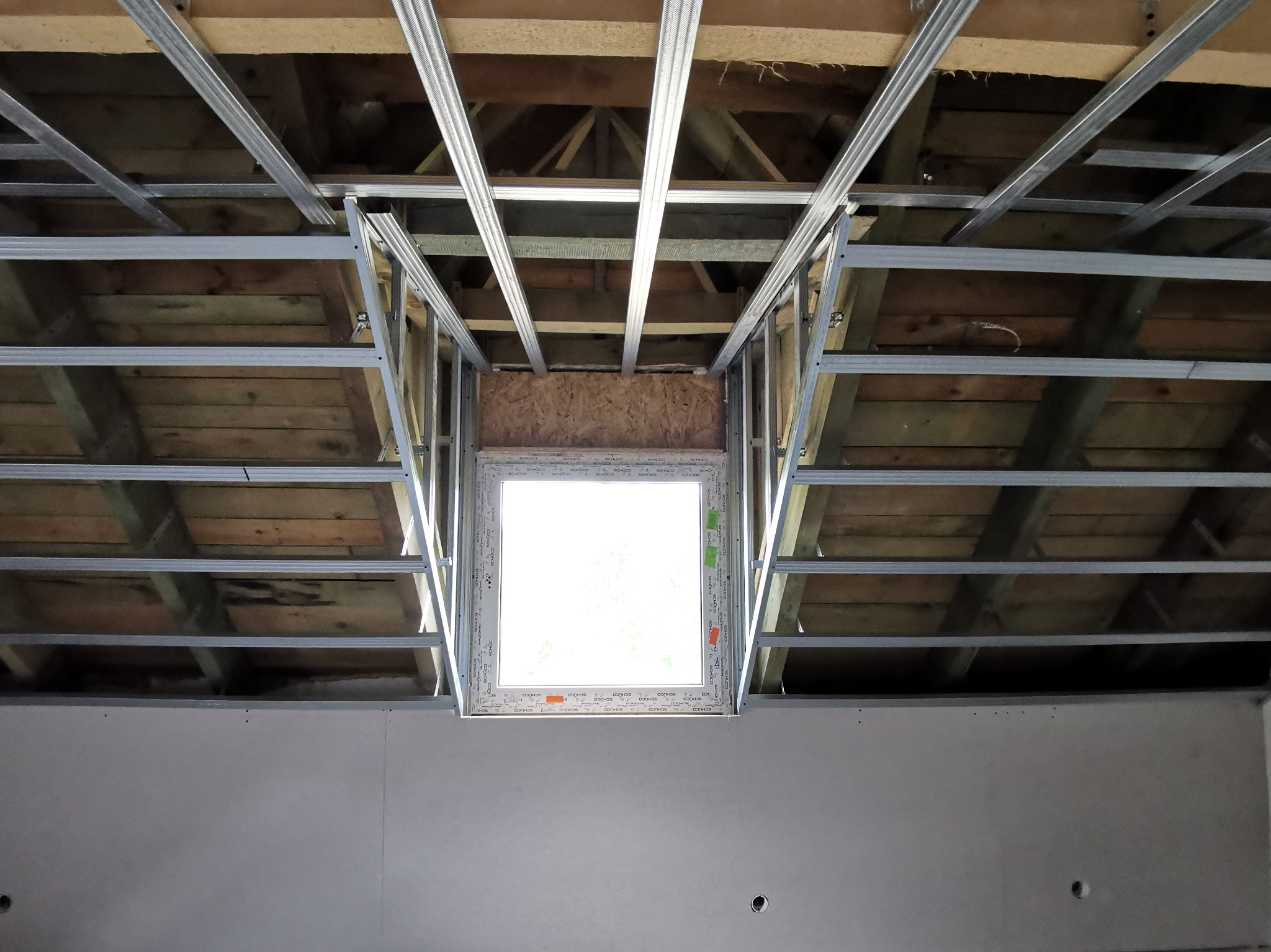 Montaż metalowej konstrukcji sufitu podwieszanego z oknem dachowym, widoczne drewniane elementy konstrukcyjne dachu i częściowo wykończona ściana.