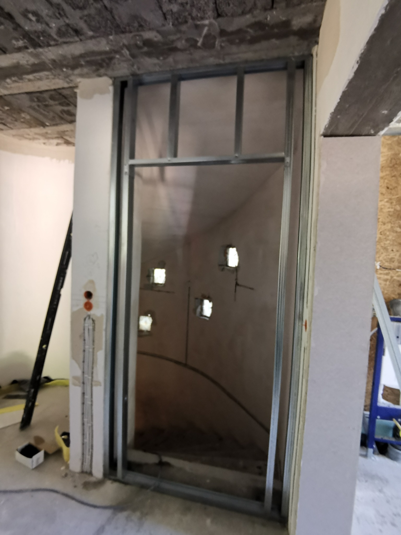 Metalowa konstrukcja ścianki działowej w trakcie montażu, widoczne przygotowane otwory na instalację elektryczną i surowy strop betonowy.