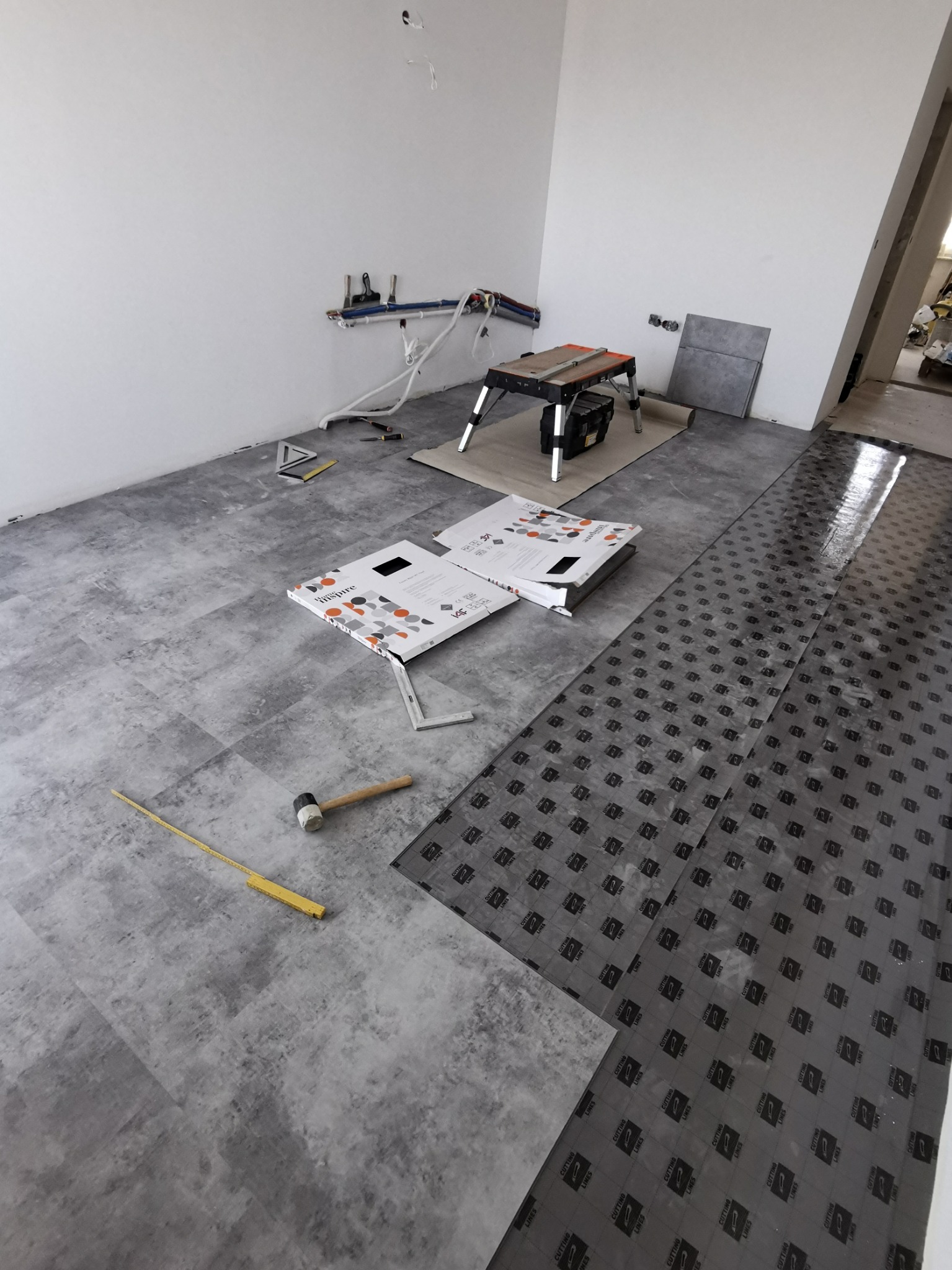 Proces układania paneli podłogowych imitujących beton, widoczna podkładka z nadrukowaną siatką do cięcia, młotek gumowy, miara składana i kartony po panelach.