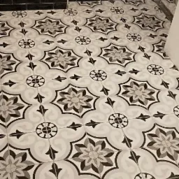 Ułożona podłoga z wzorzystych, szaro-białych płytek ceramicznych w łazience, z fragmentem ściany wyłożonej czarnymi, błyszczącymi płytkami typu metro.