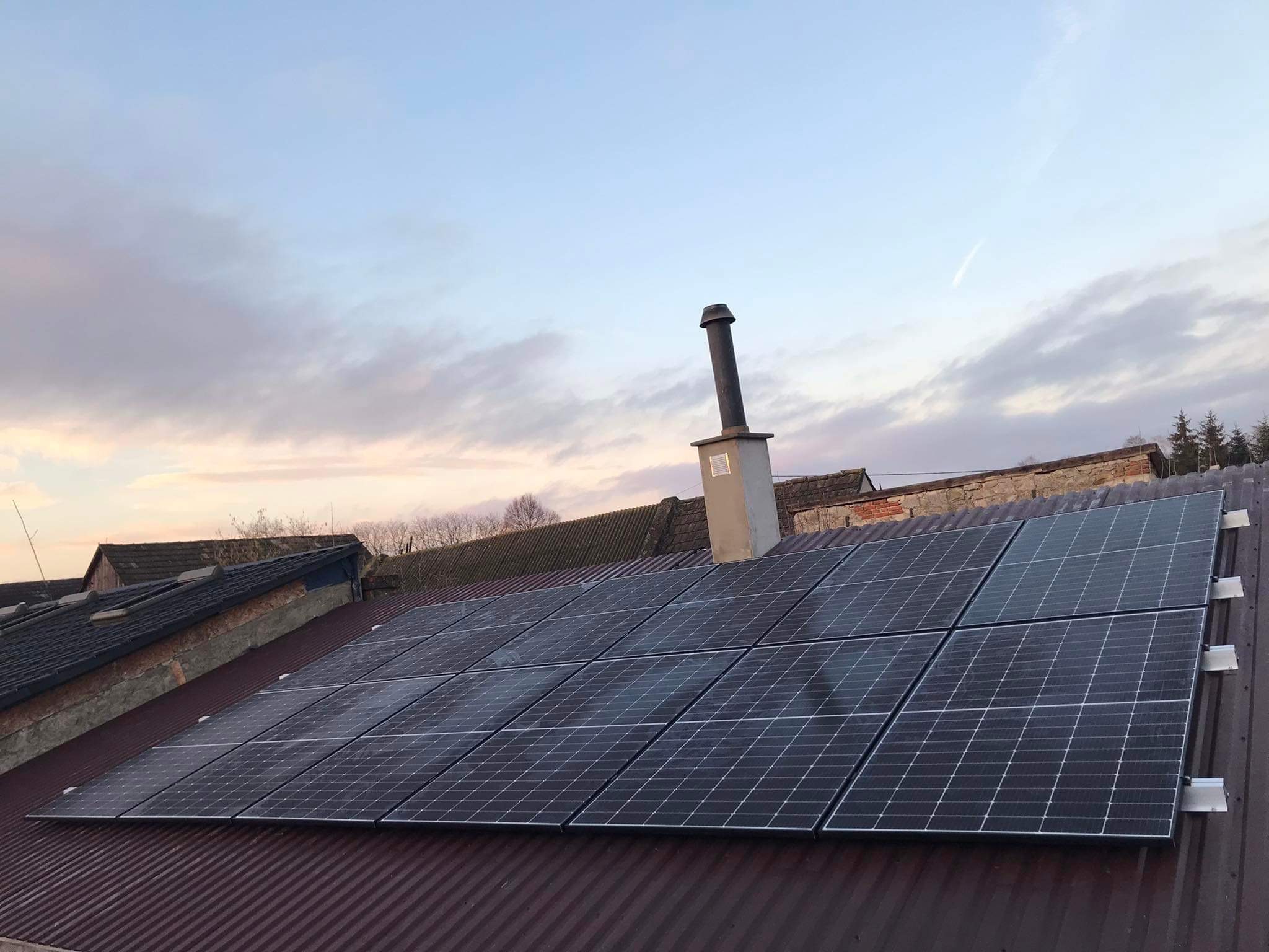 Panele słoneczne zamontowane na falistym, metalowym dachu budynku z kominem, sfotografowane pod wieczornym niebem z delikatnymi obłokami.