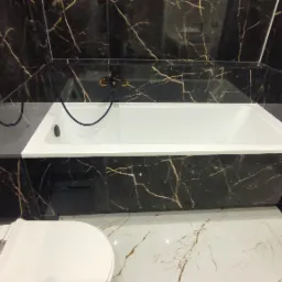 Biała wanna obudowana czarnymi płytkami imitującymi marmur ze złotymi żyłami, widok z góry na fragment toalety i podłogę z podobnym wzorem płytek.