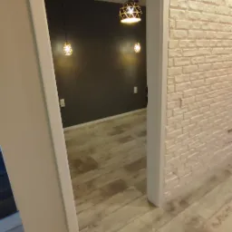 Wnętrze mieszkania po remoncie: ściana z białej cegły, drewnopodobna podłoga i nowoczesne lampy wiszące widziane przez framugę drzwi.