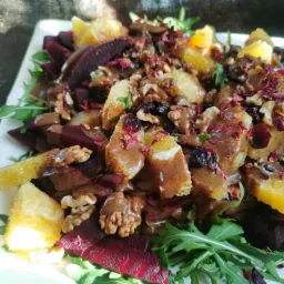 Kompozycja sałatki z burak&oacute;w, pomarańczy, rukoli, orzech&oacute;w włoskich, suszonej żurawiny, polana sosem tahini i posypana płatkami r&oacute;ż, podana na białym kwadratowym talerzu.