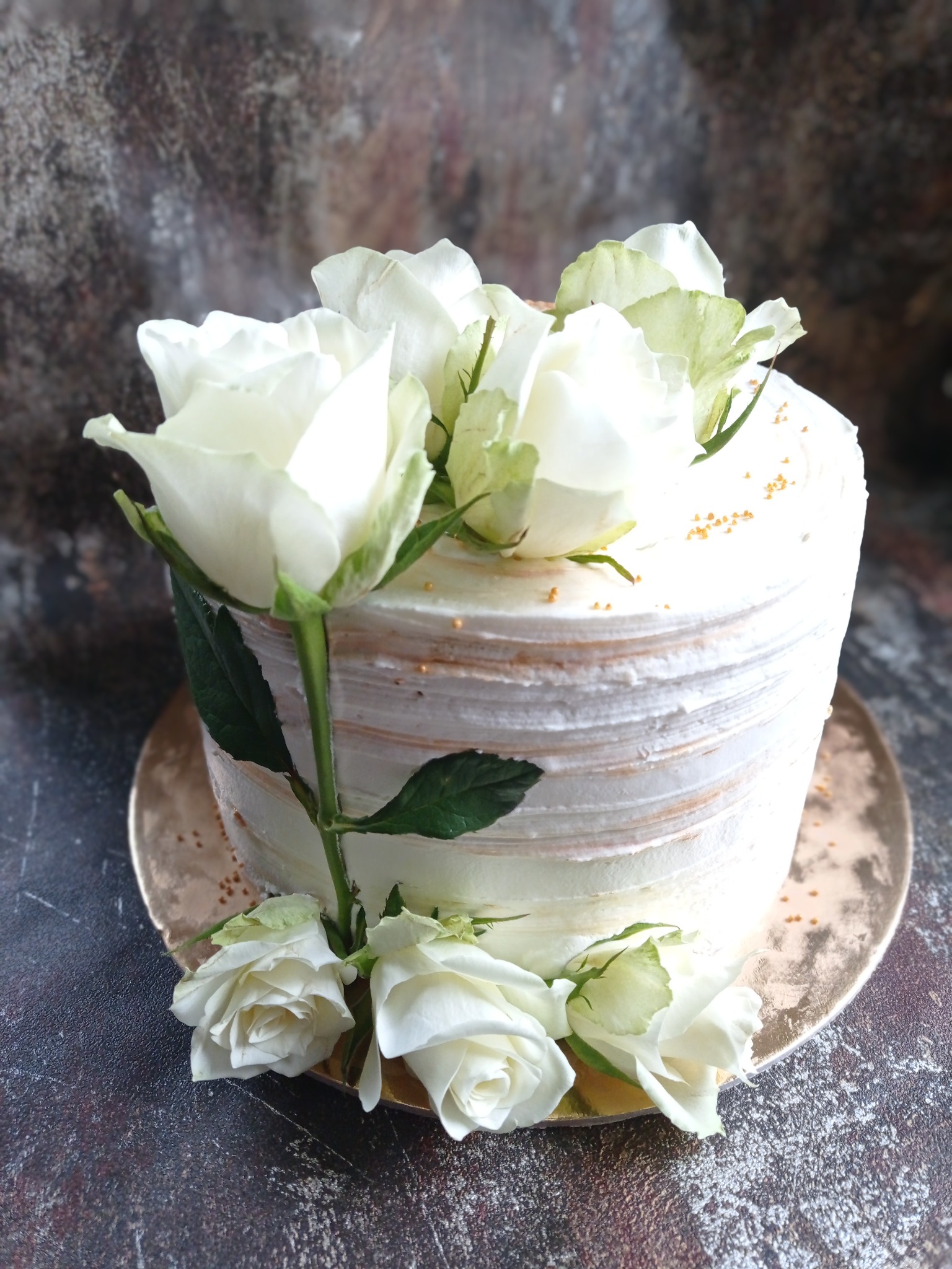 Tort udekorowany białymi różami i złotymi posypkami na złotym talerzu, na tle ciemnej, teksturowanej powierzchni.