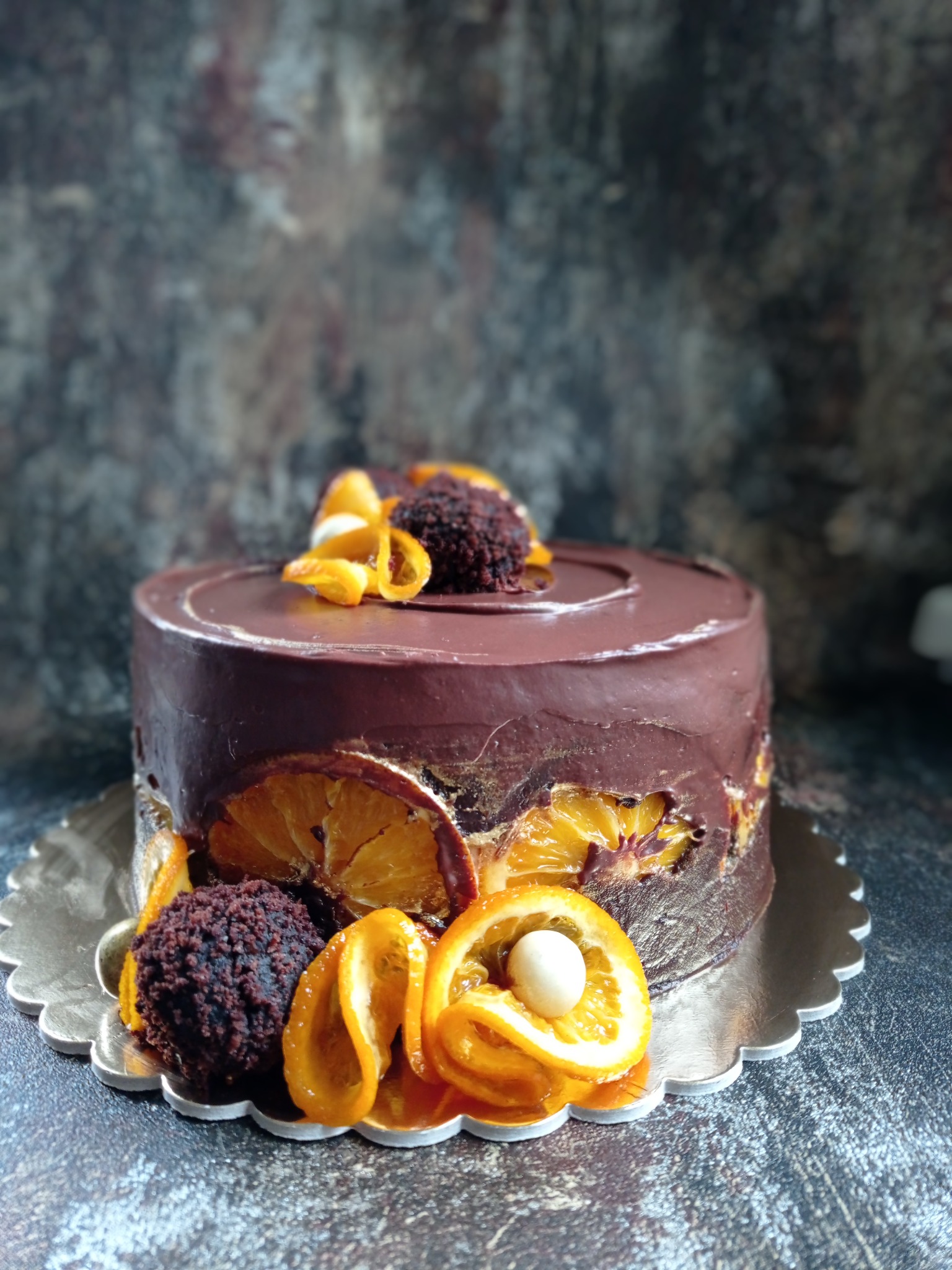 Ciemny tort czekoladowy udekorowany kandyzowanymi pomarańczami, kulkami czekoladowymi i kremem na srebrnej podkładce.