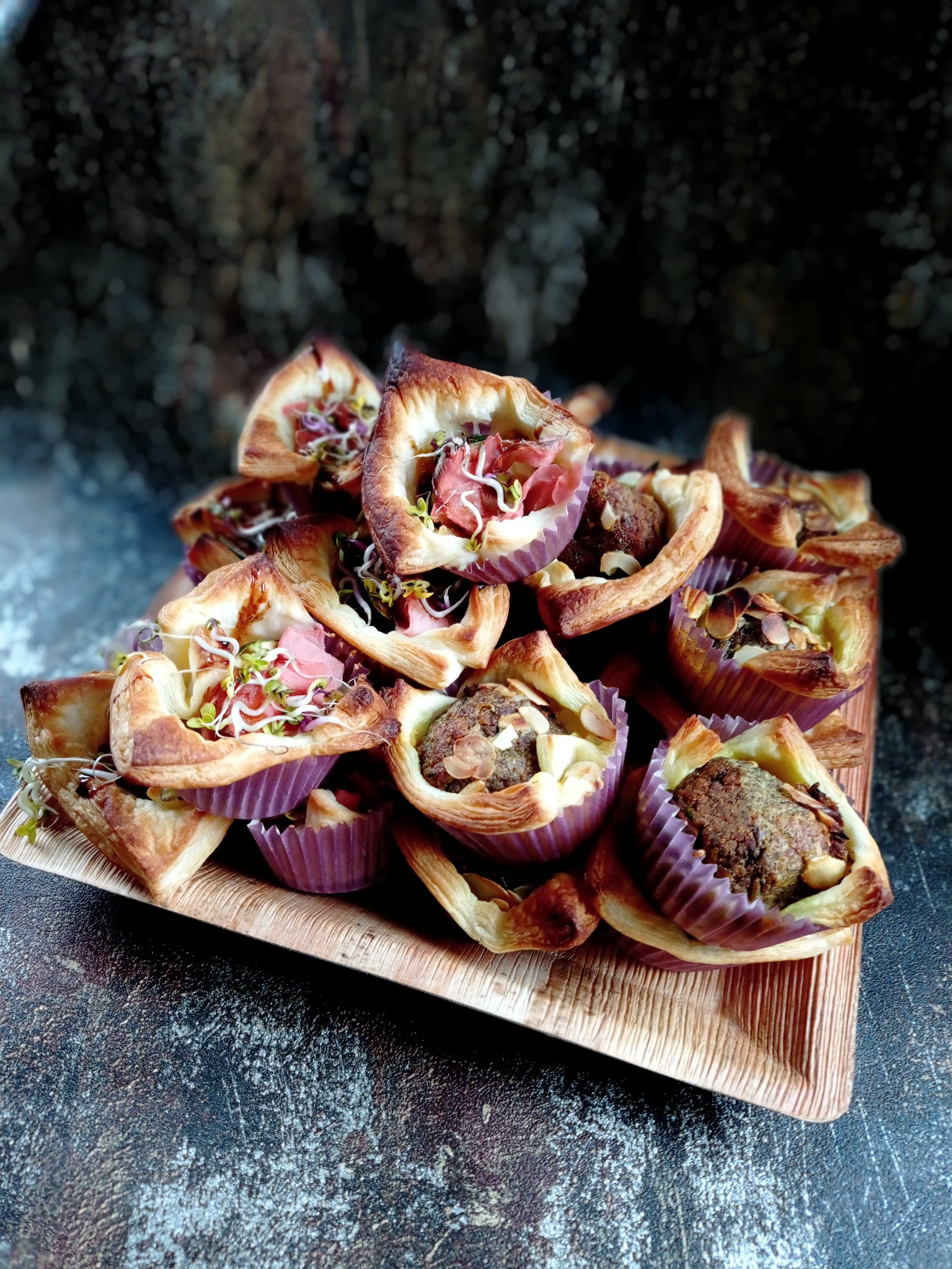 Kruche babeczki z różnymi nadzieniami, część z szynką i kiełkami, część z farszem mięsnym i migdałami, ułożone na drewnianej tacy na ciemnym tle.