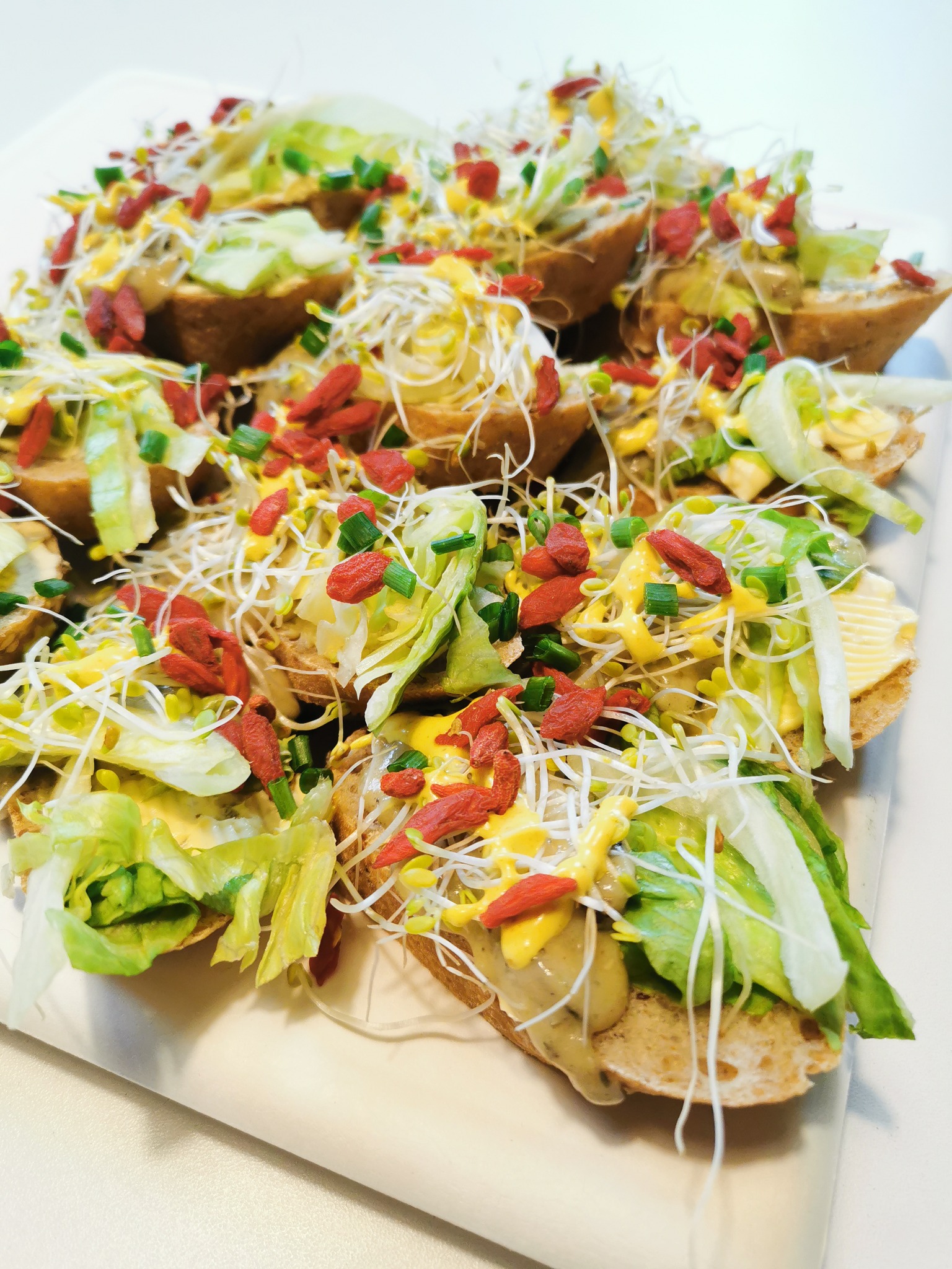 Mini kanapeczki na jasnym pieczywie z sałatą, kiełkami, sosem i owocami goji, ułożone na białym talerzu.