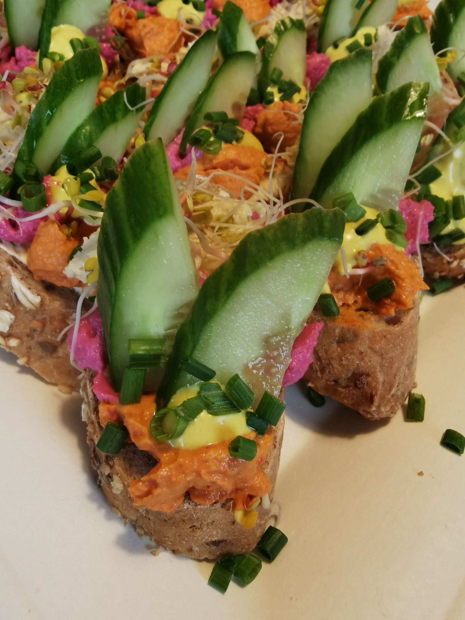 Kanapki z chleba wieloziarnistego z różnymi pastami, ogórkiem i szczypiorkiem, ułożone na białym talerzu.